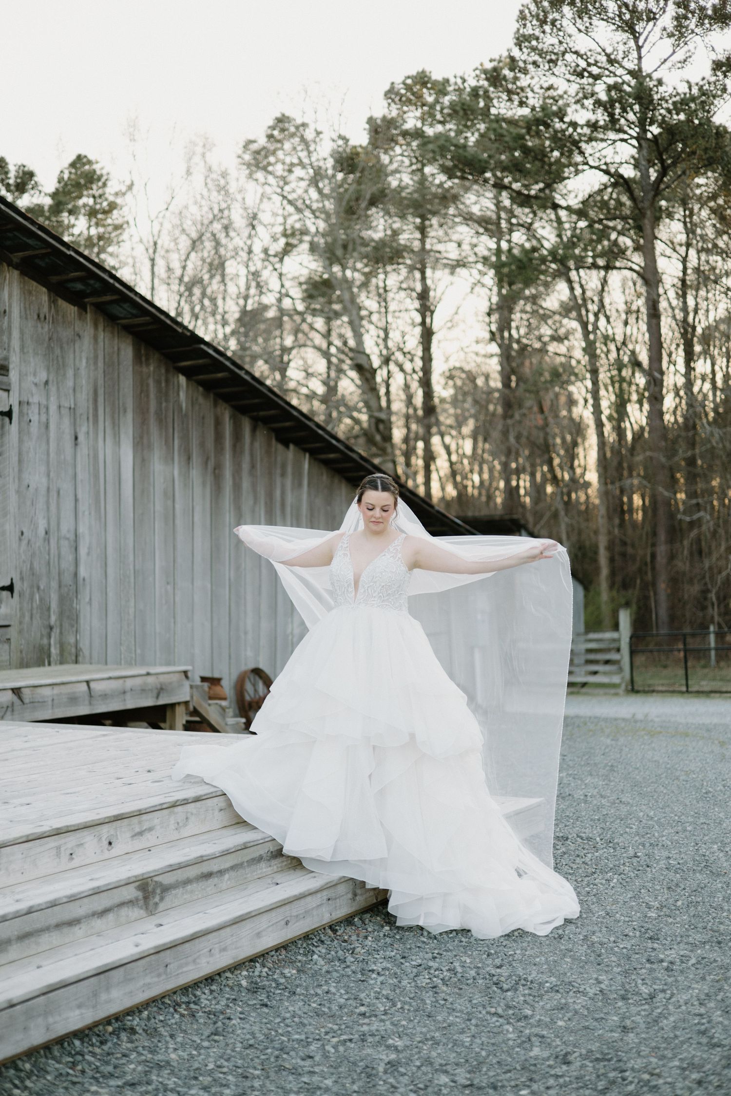 A flowy white wedding gown flows gracefully on the wooden steps of a rustic barn surrounded by tall trees at dusk.