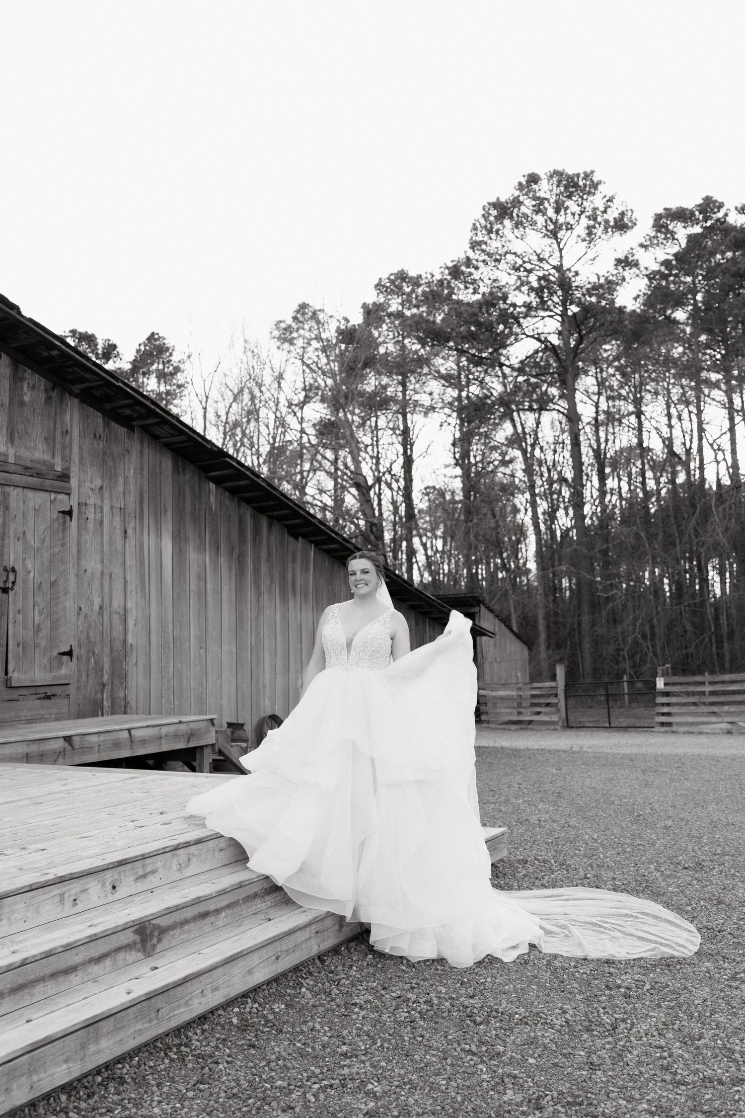 A flowing white wedding dress photographed in front of a rustic wooden barn structure in a dramatic outdoor setting.