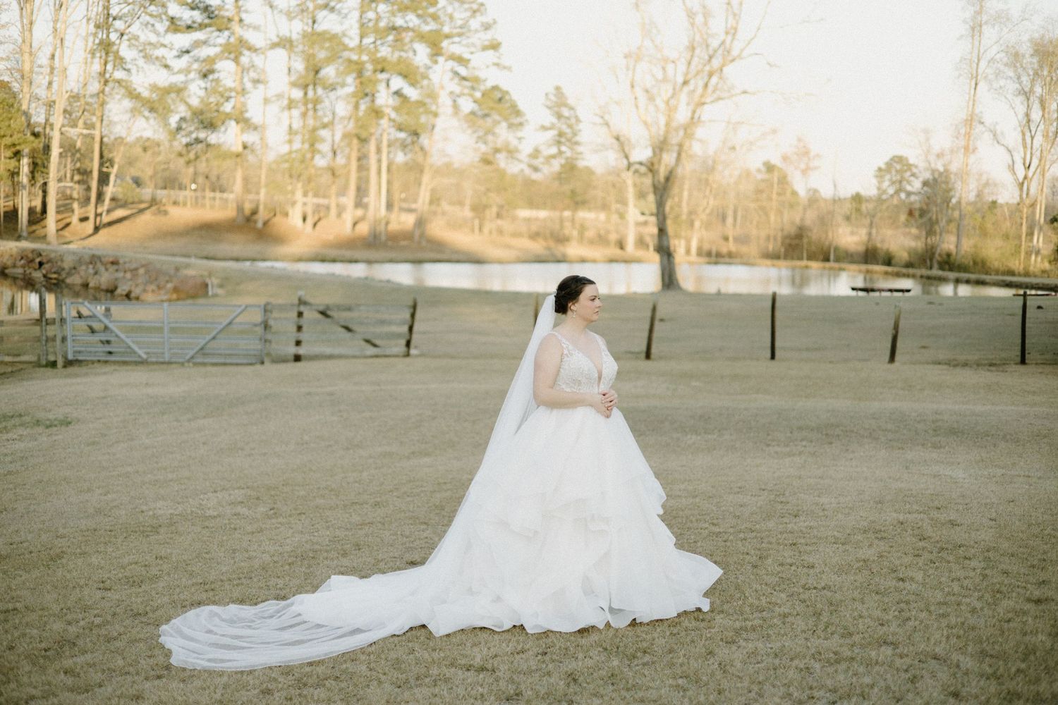 A bride in a flowing white wedding gown and veil stands on a grassy field at sunset with winter trees in the background.