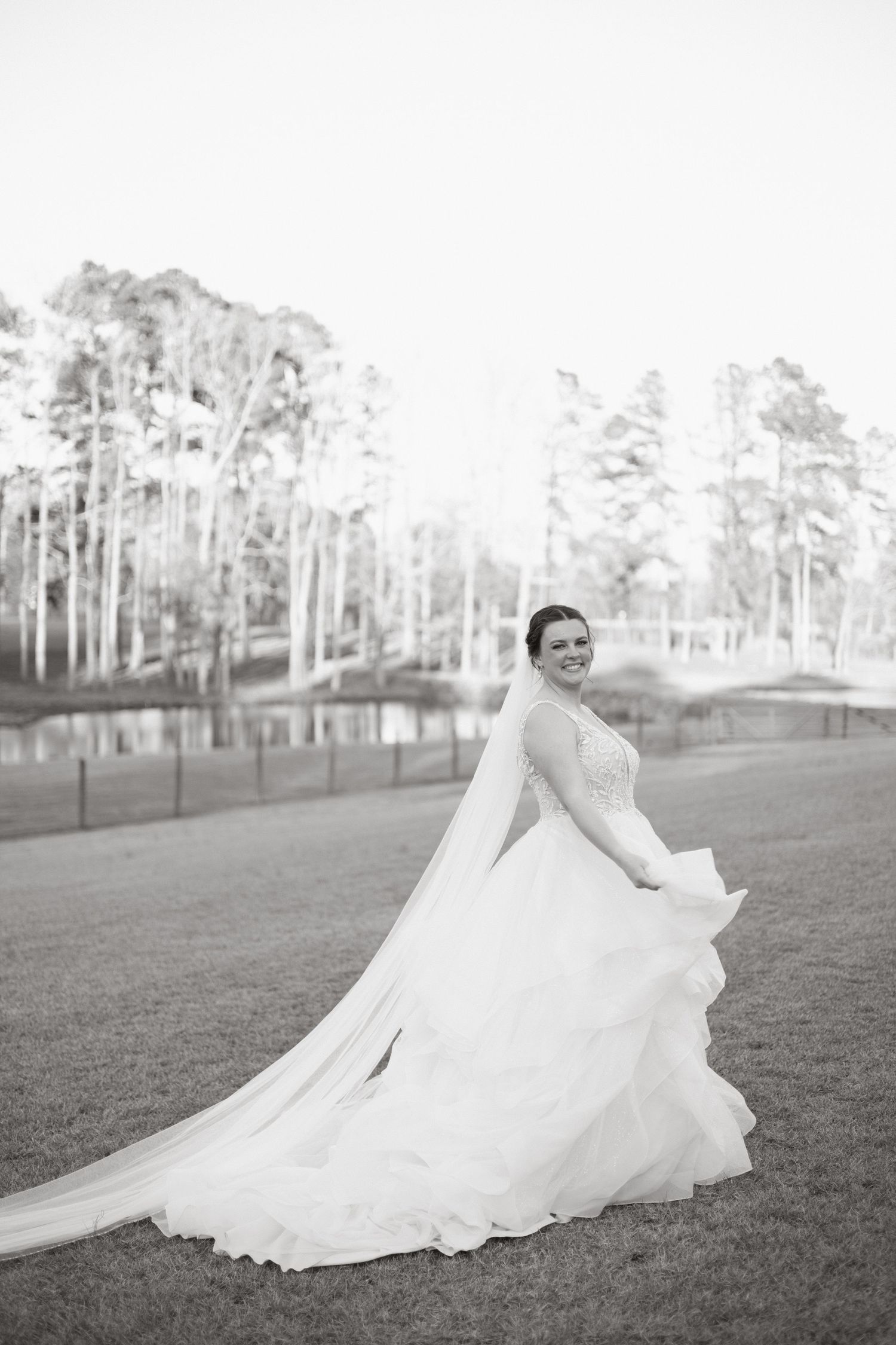 A bride in an elegant white wedding dress with a long train twirls on a grassy lawn near tall pine trees.