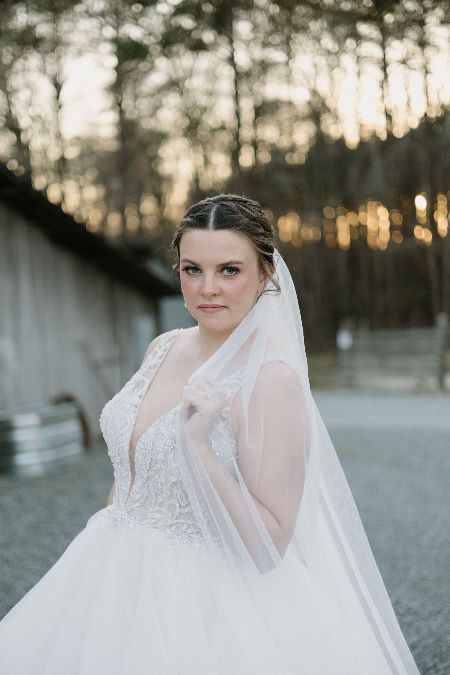 Bride wearing a white lace wedding gown with flowing veil poses at sunset with trees in background.