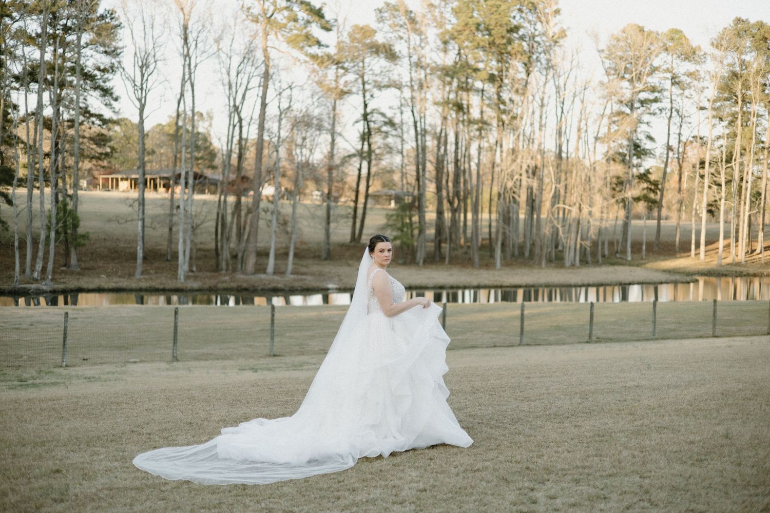 A flowing white wedding gown cascades across a rustic outdoor setting surrounded by tall trees at sunset.
