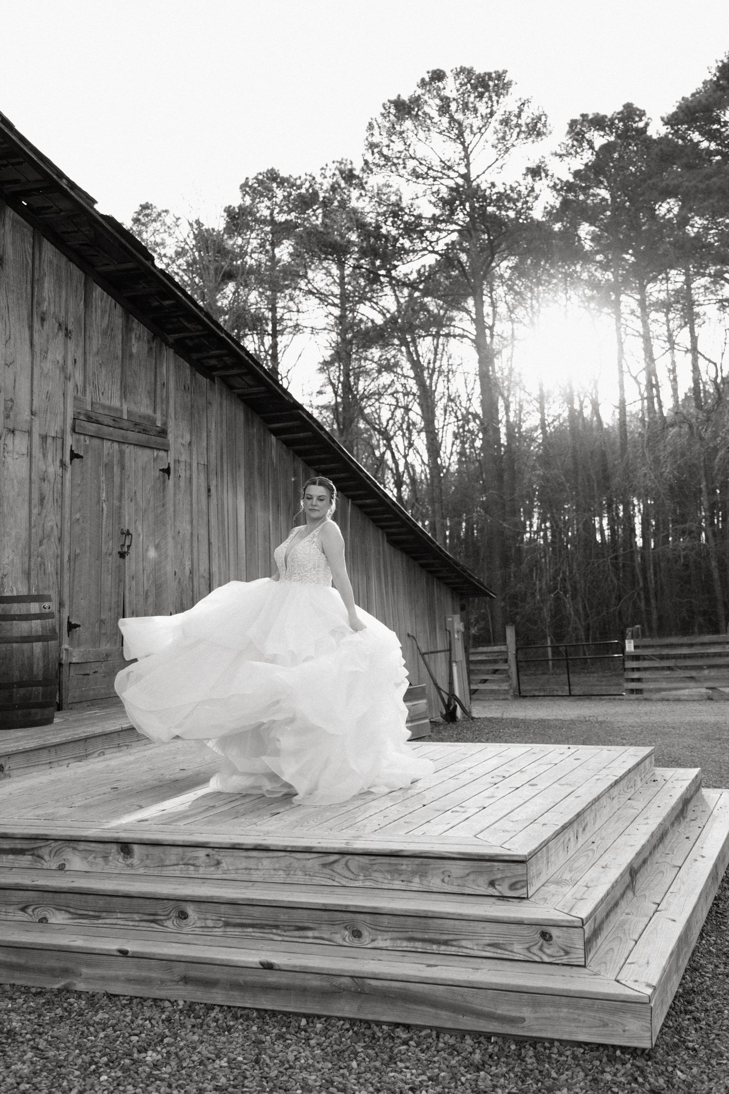 A flowing white wedding dress billows dramatically on wooden steps outside a rustic barn with sunlit pine trees in background.