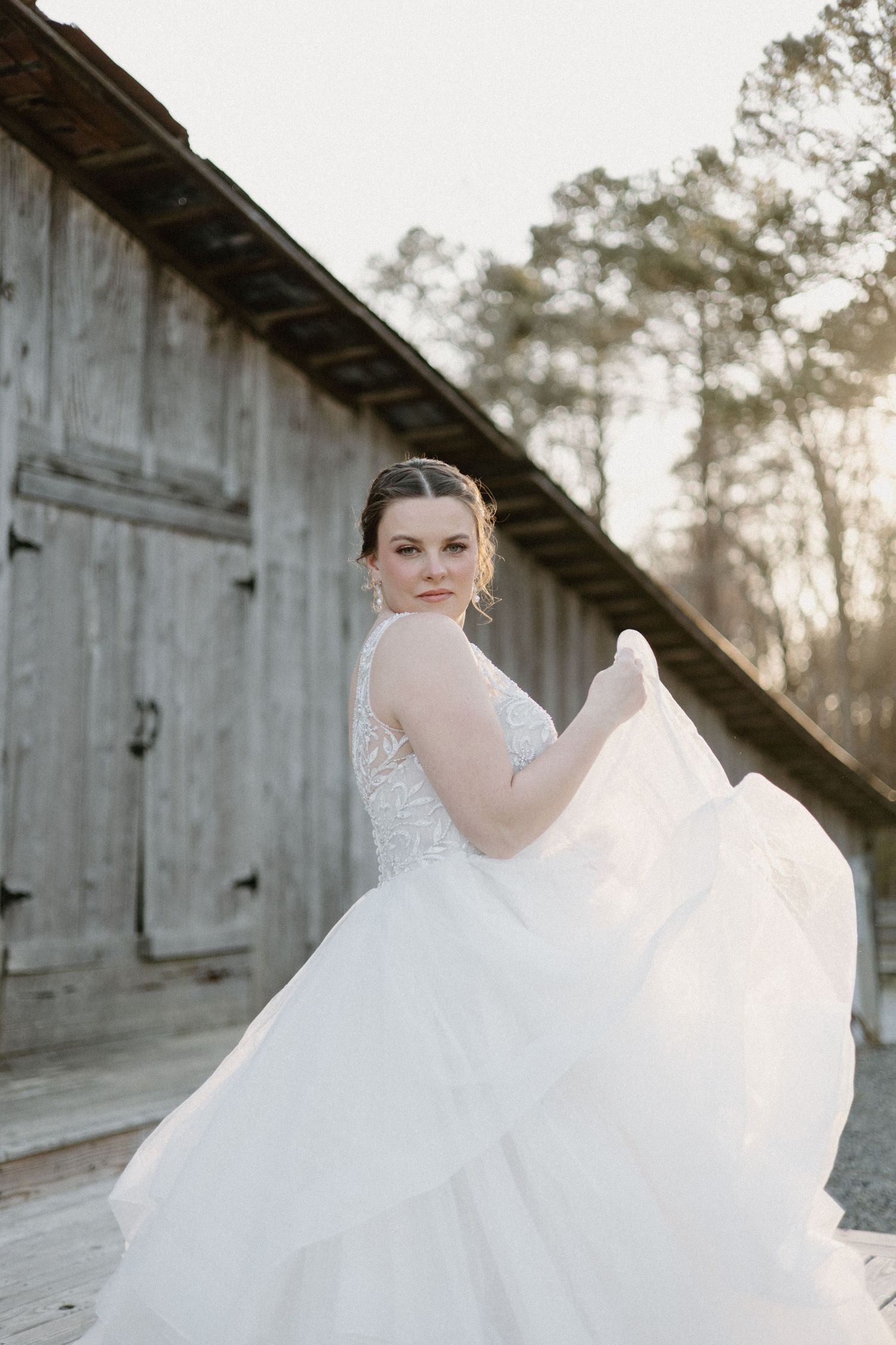 A flowing white wedding gown swirls gracefully against a rustic barn backdrop at sunset.