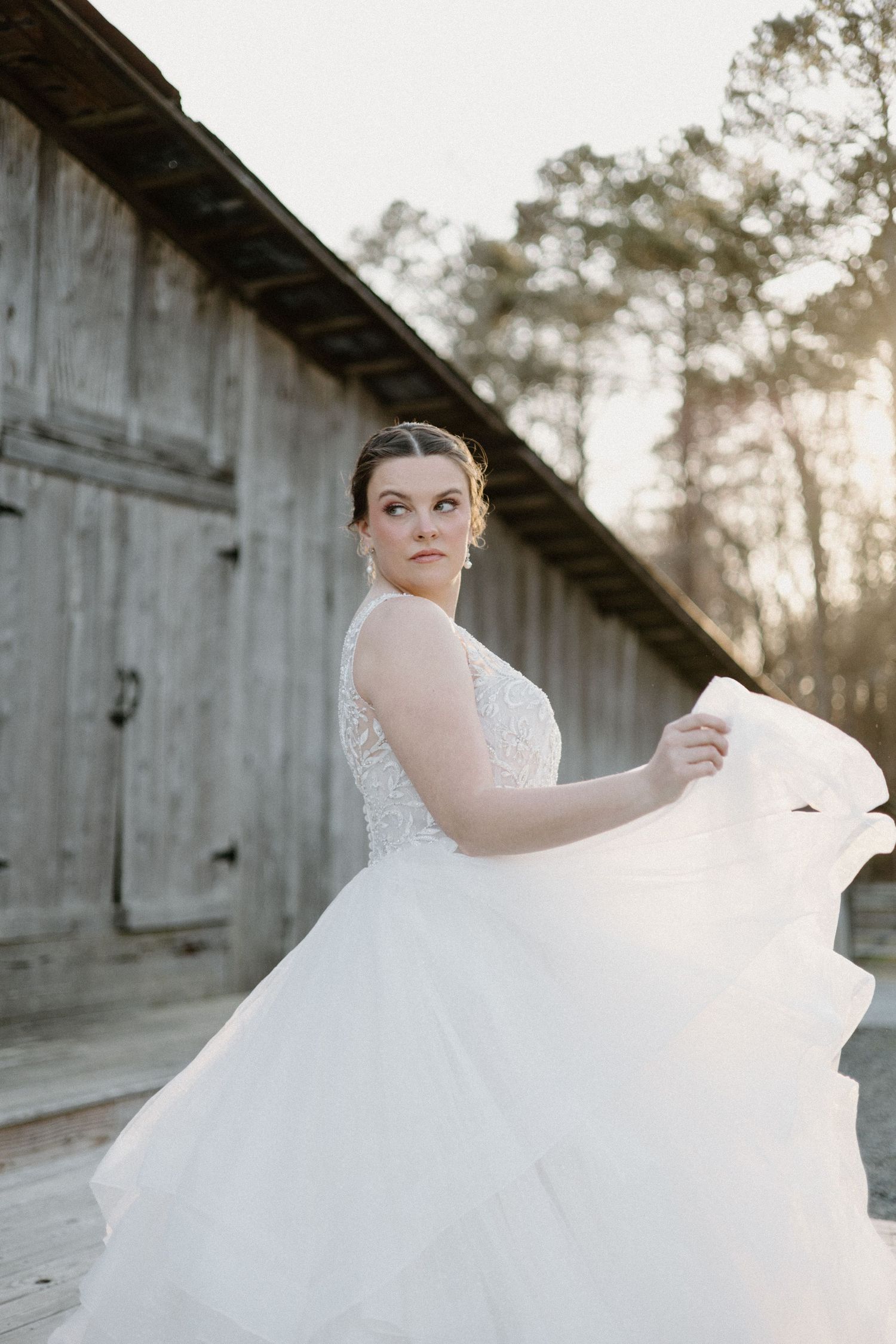 A person in a white lace wedding gown twirls gracefully against a rustic barn backdrop during sunset.