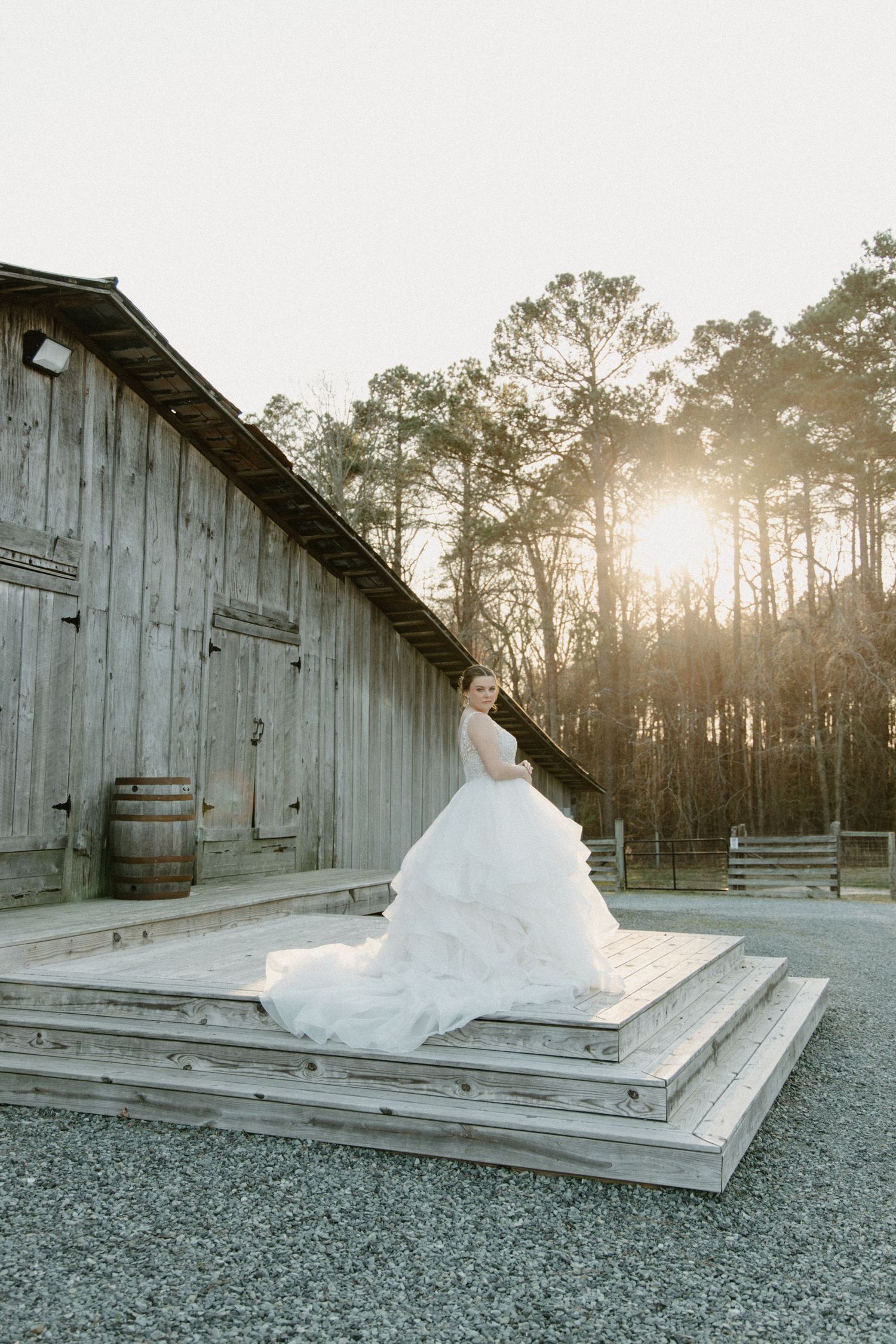 A minimalist white sculptural installation rests on a wooden platform against a rustic barn at sunset with tall pines beyond.