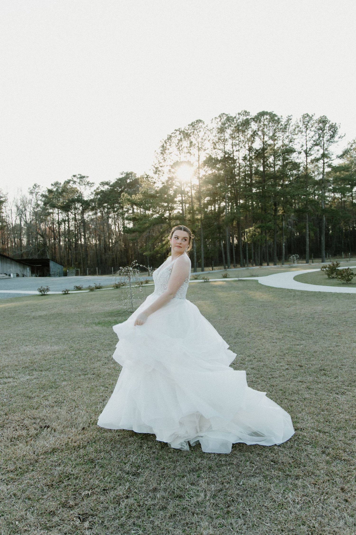A bride in a flowing white gown twirls gracefully in a golden-lit field with tall pine trees in the background.