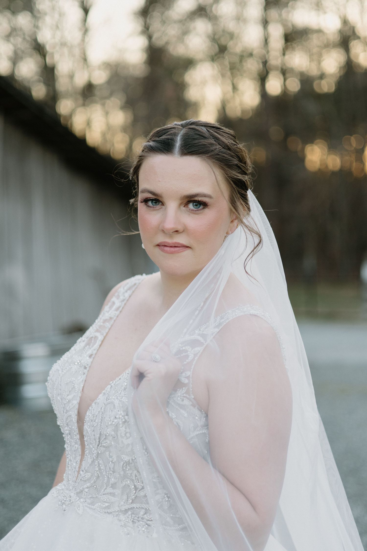Close-up portrait of a bride wearing a delicate white lace wedding gown and veil against a blurred outdoor background.