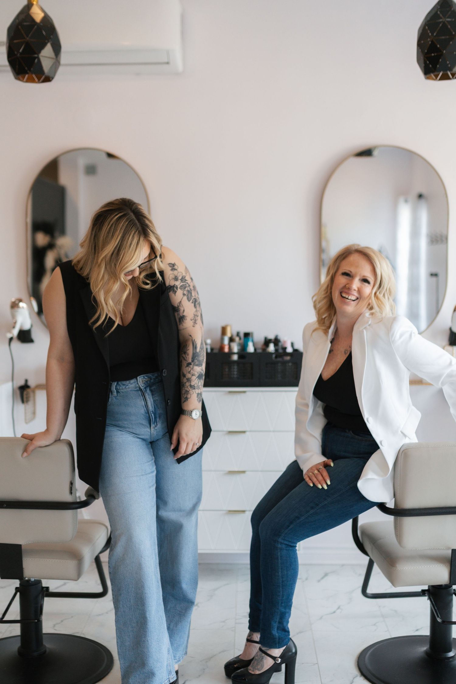 Two women laughing together in modern hair salon with oval mirrors and styling chairs.