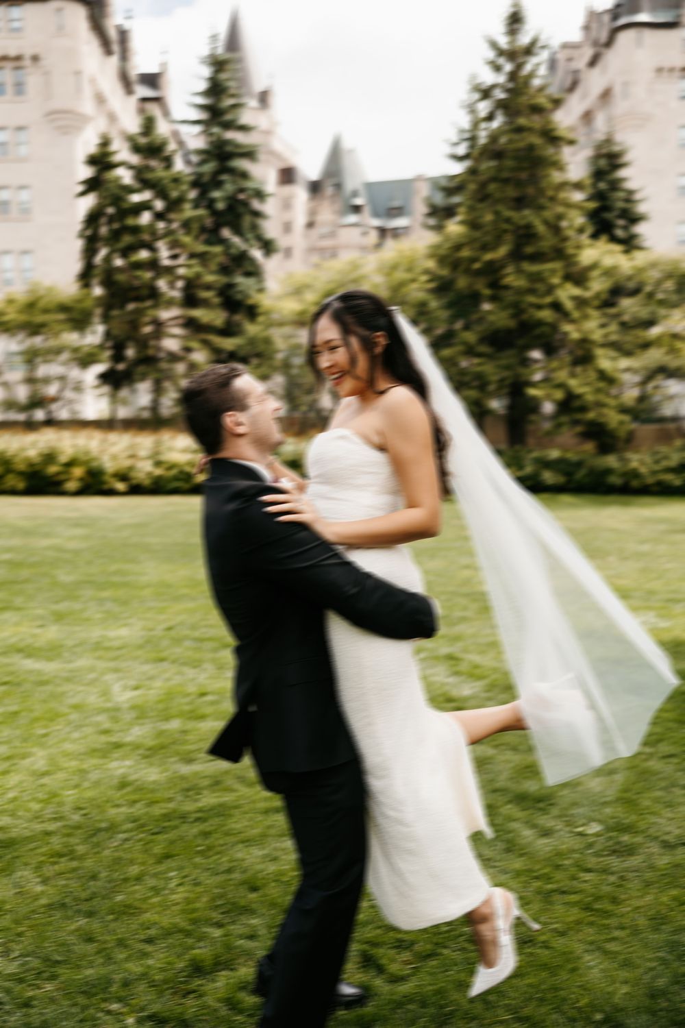 A romantic wedding couple dances and spins together on a grassy lawn with evergreen trees in the background.