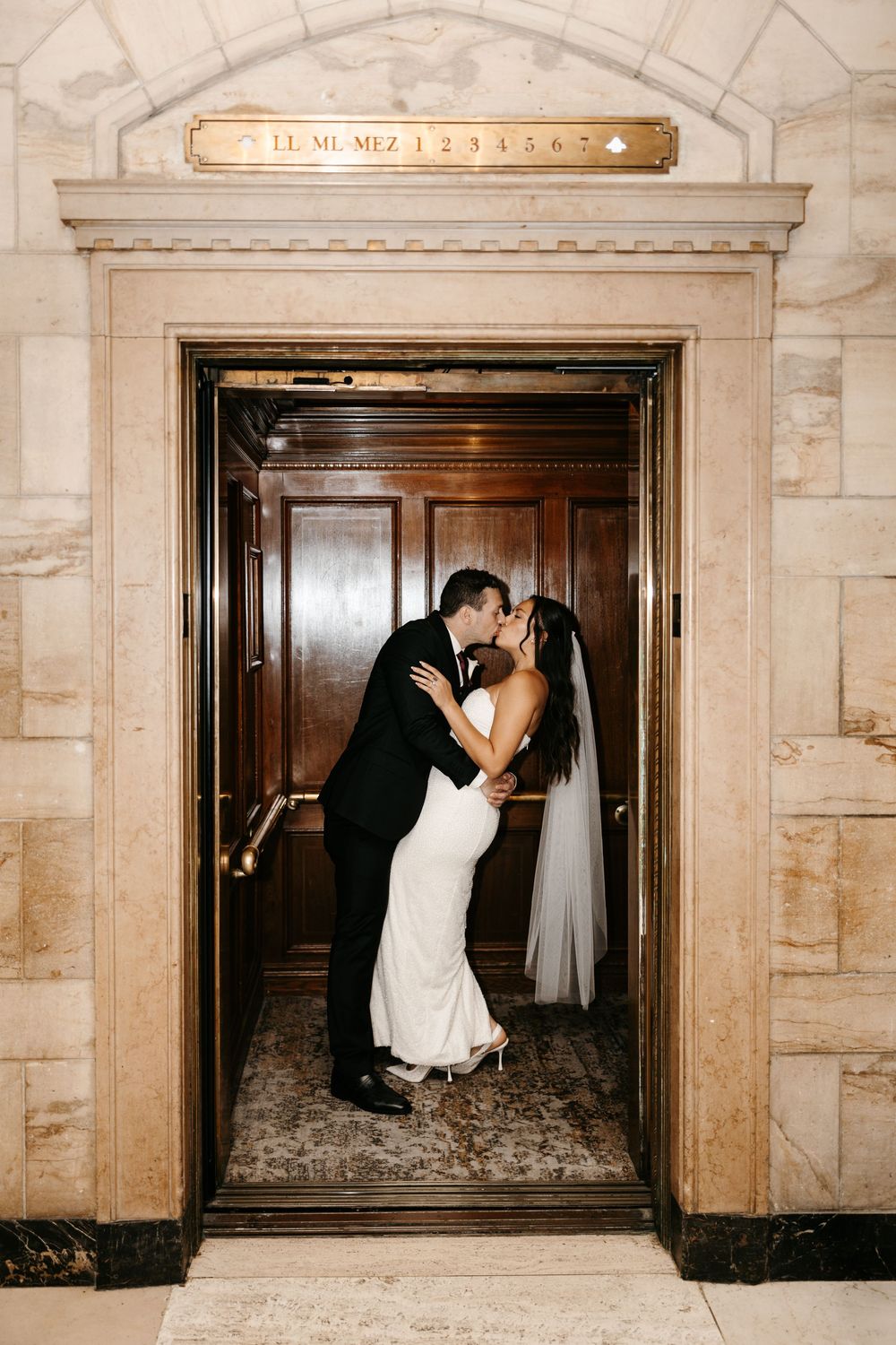 Romantic wedding couple dips and kisses in front of vintage elevator doors in classic marble hallway.