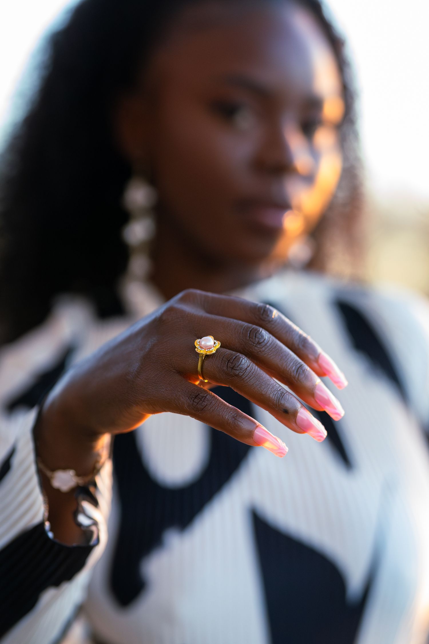 Close-up of a hand wearing coral nail polish and a gold statement ring against a black and white patterned background.