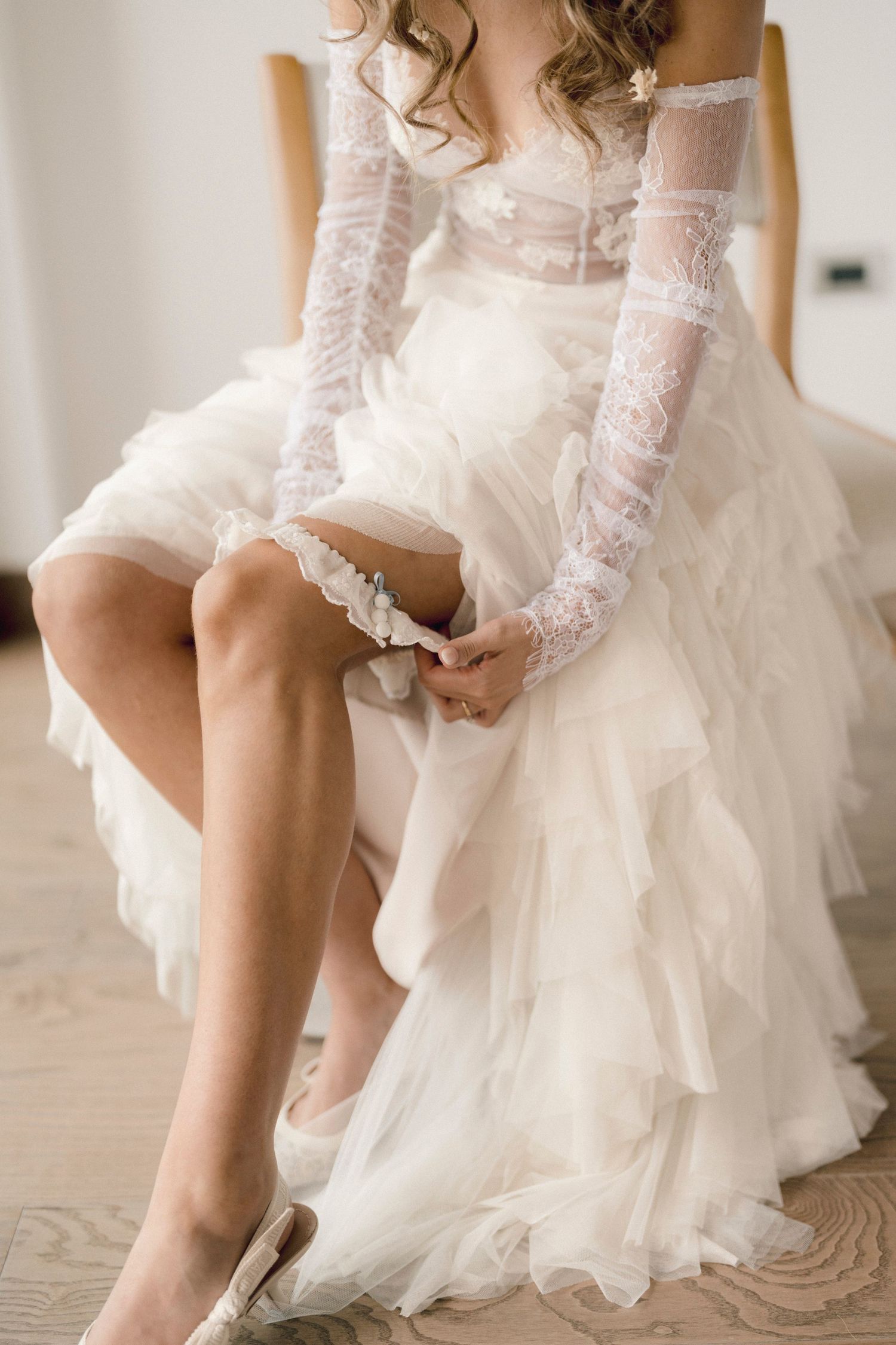Bride in white lace wedding gown adjusts delicate garter on leg while sitting in elegant bridal suite.