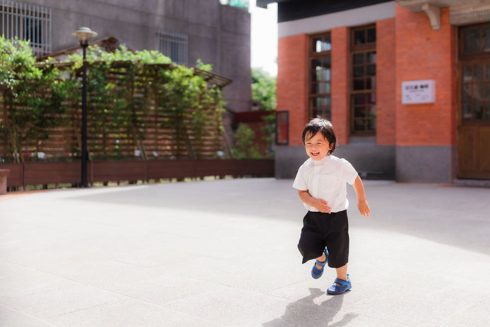 A small child runs playfully in an open courtyard space with greenery in the background.