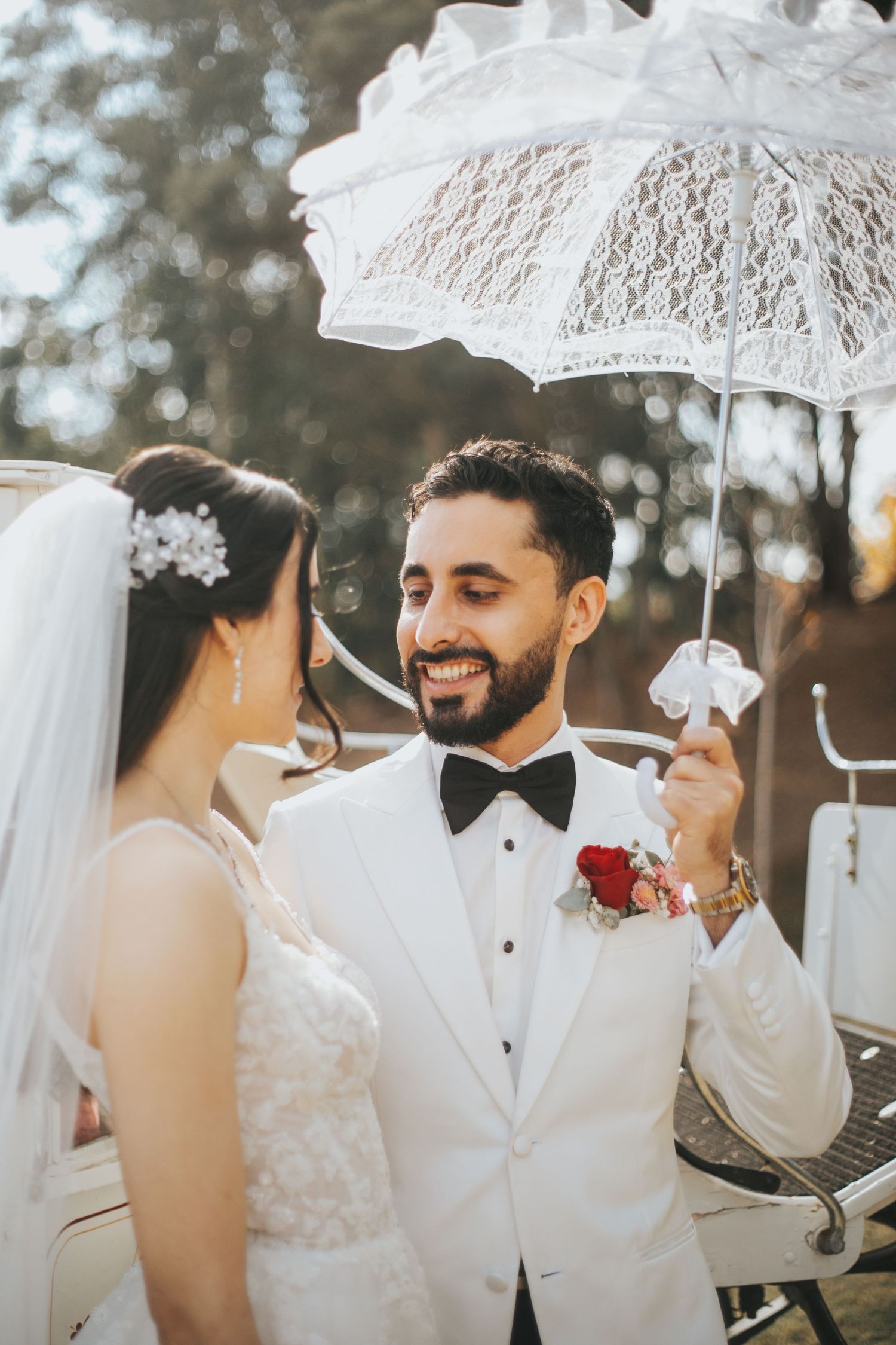 Smiling groom in white tuxedo holds lace umbrella beside bride in wedding dress outdoors.