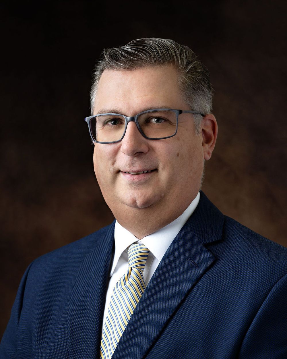 Professional headshot of someone in a navy suit and striped tie against dark background.
