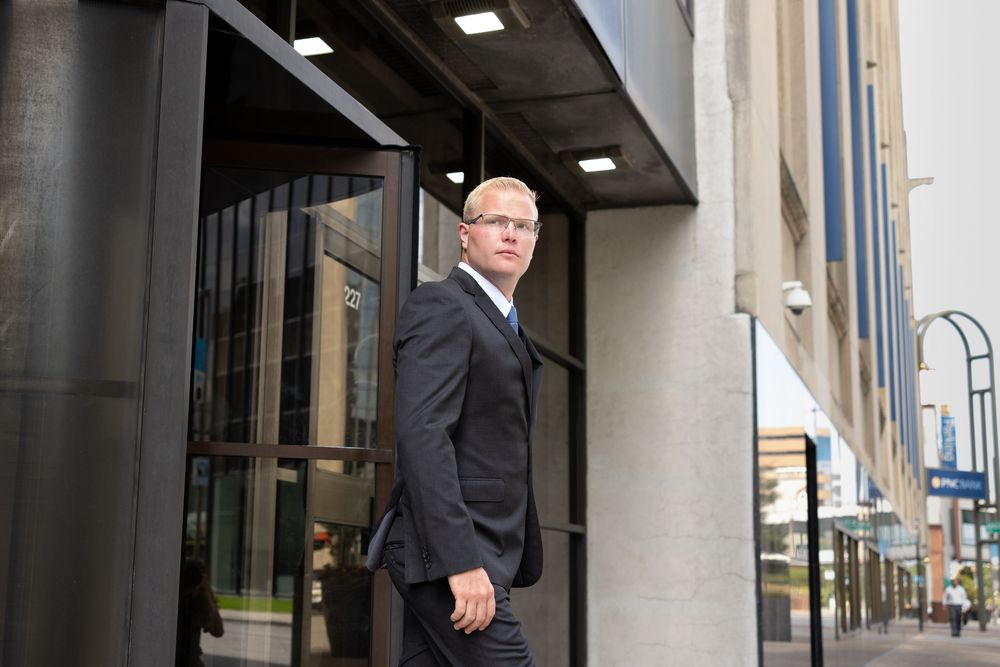 Business professionals in dark suits walk through a modern office building corridor with glass walls and overhead lighting.