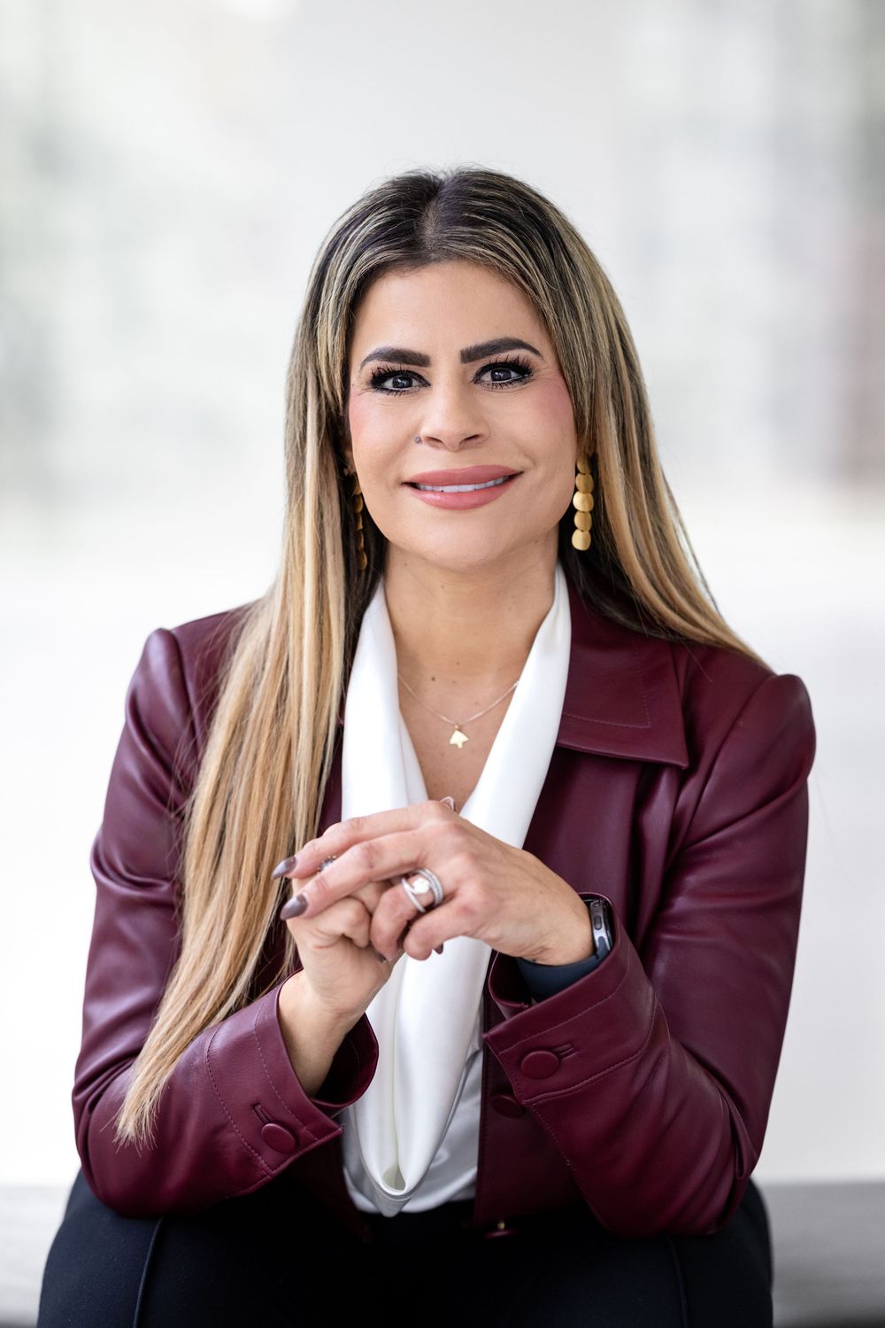Professional headshot of someone in a burgundy blazer and white blouse with long straight hair and gold earrings.