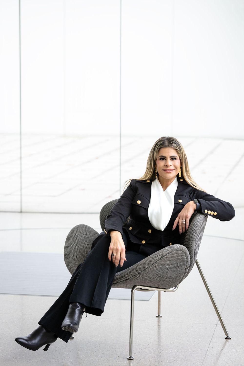 Professional wearing a black blazer and white blouse sits confidently in a modern grey chair against white background.