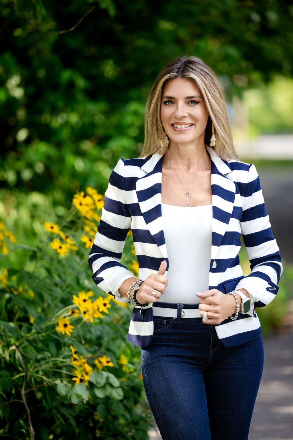 A person wearing a navy and white striped blazer with jeans poses outdoors near yellow flowers in a garden setting.
