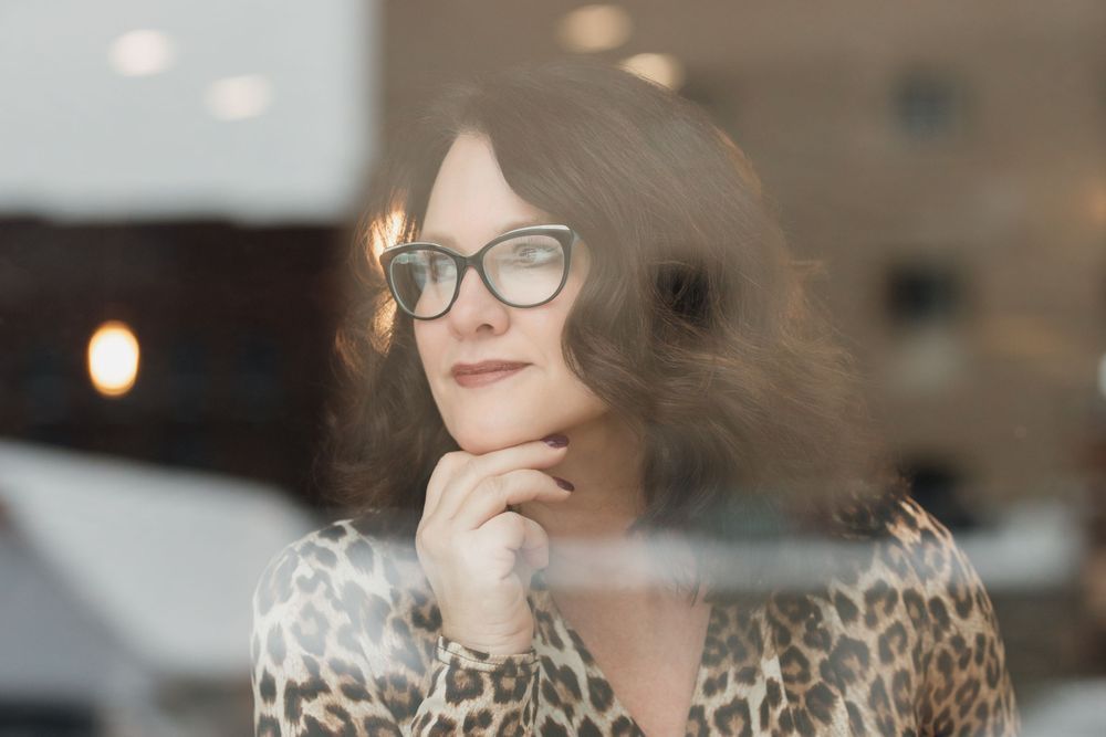Through glass window, person with glasses and leopard print top rests chin on hand while looking away thoughtfully.