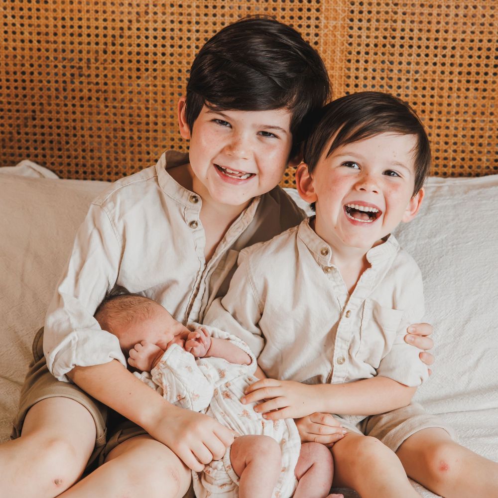 Two smiling siblings sitting together while holding their newborn baby during a natural family photoshoot.
