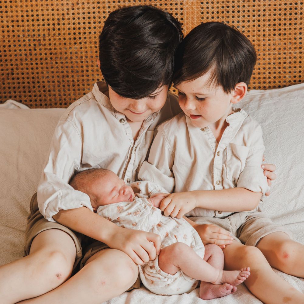 Two young siblings gently holding their newborn baby on a bed in a warm, neutral-toned family photoshoot.