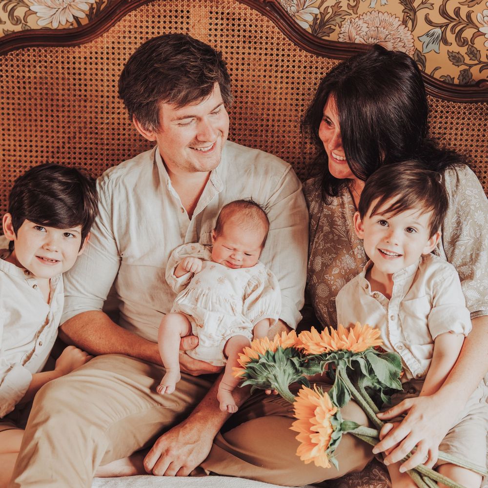 Family of 5, at home cuddled up on a bed. Dad is holding newborn baby and siblings are laughing together. Newborn home shoot.