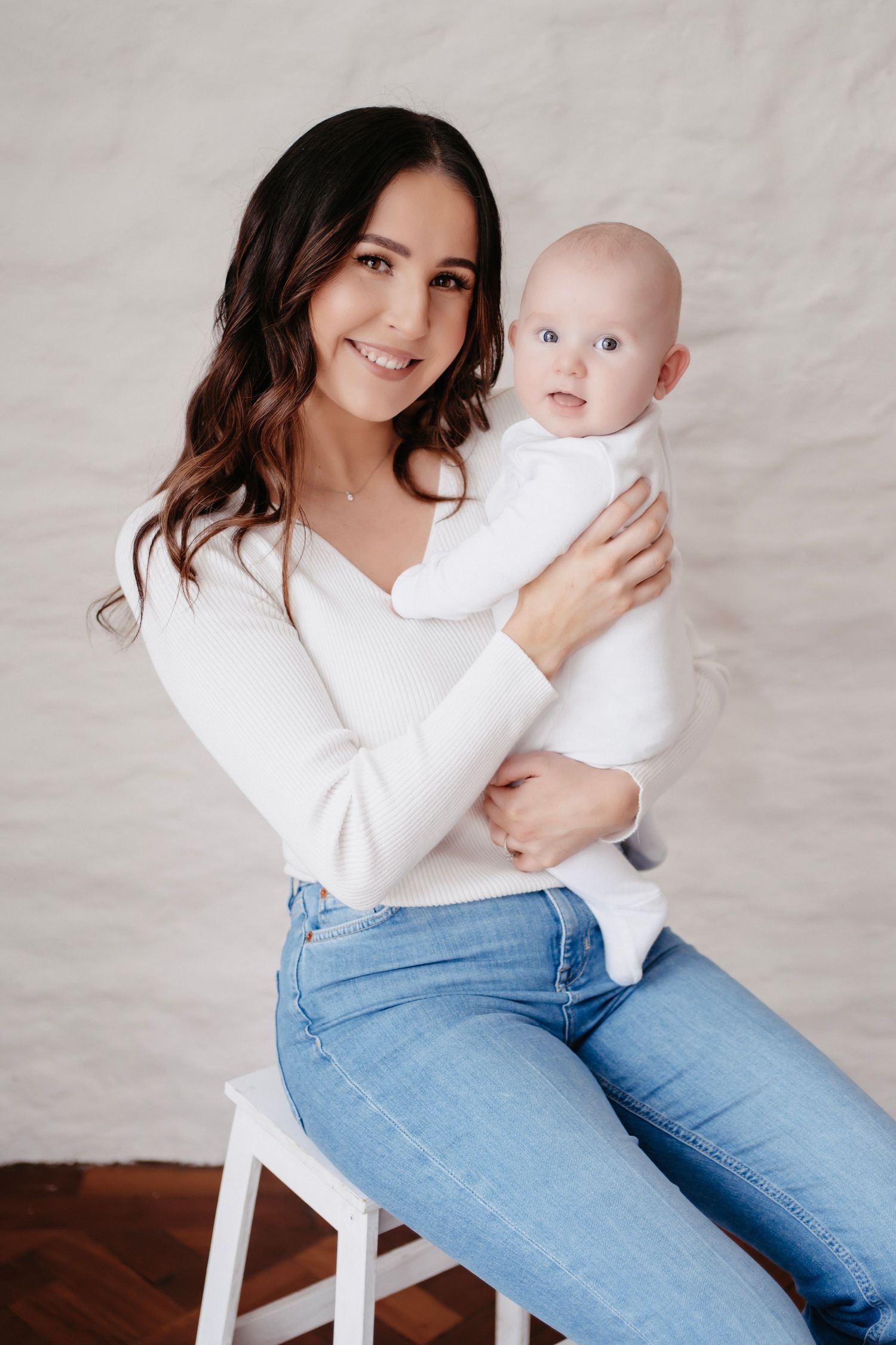 A parent in blue jeans and white sweater holds a baby in white while sitting on a white chair against a neutral backdrop.