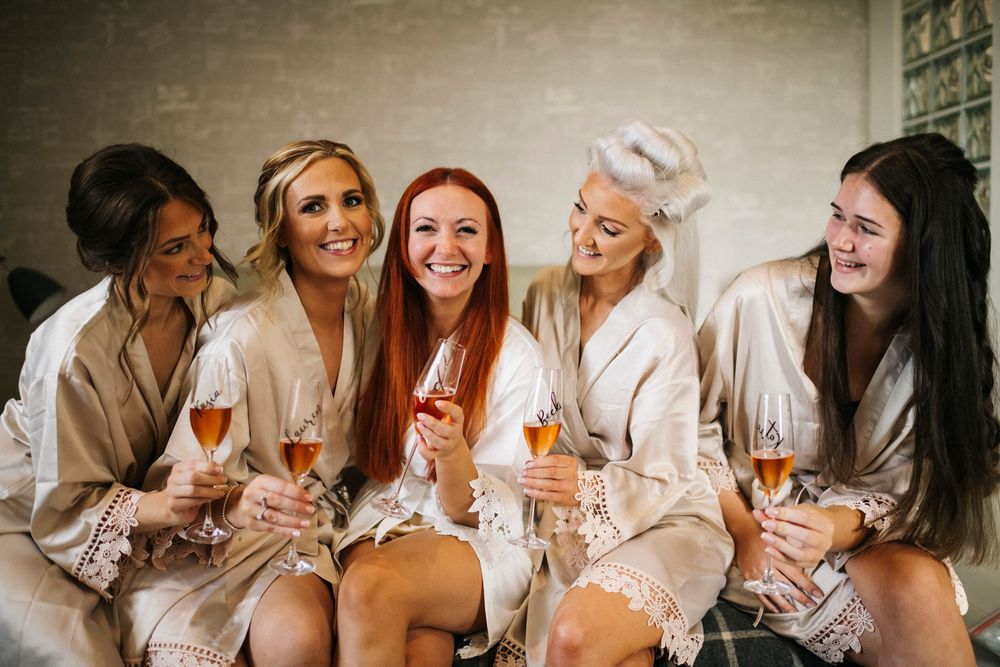 Bridesmaids in white robes toasting with wine glasses during wedding preparations.