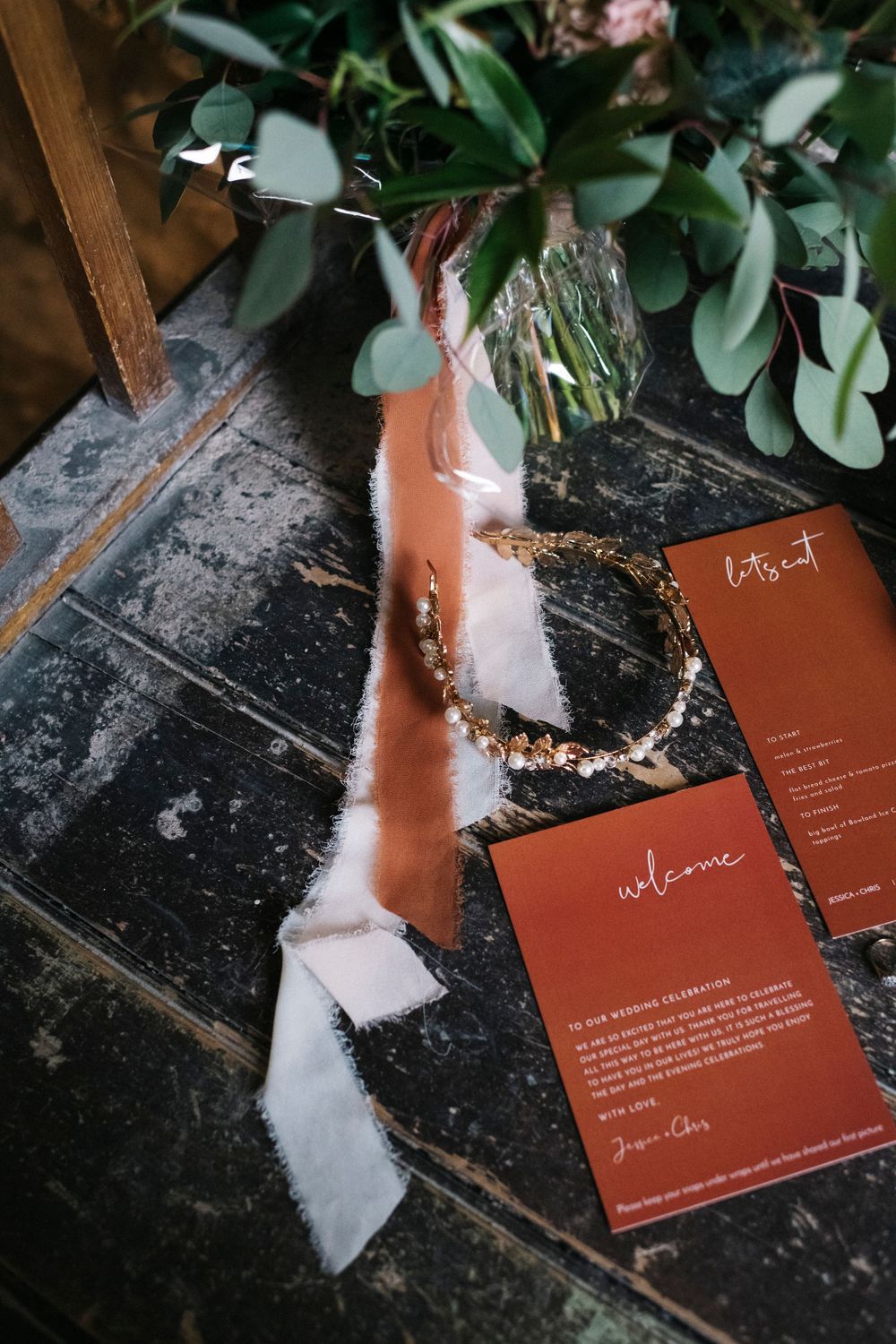 Terracotta wedding invitations with white text displayed with greenery and ribbon accents.