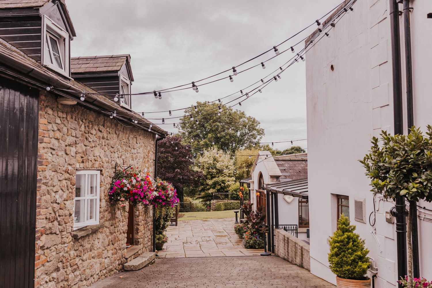 String lights hang between rustic stone buildings creating a charming outdoor walkway at a countryside venue.