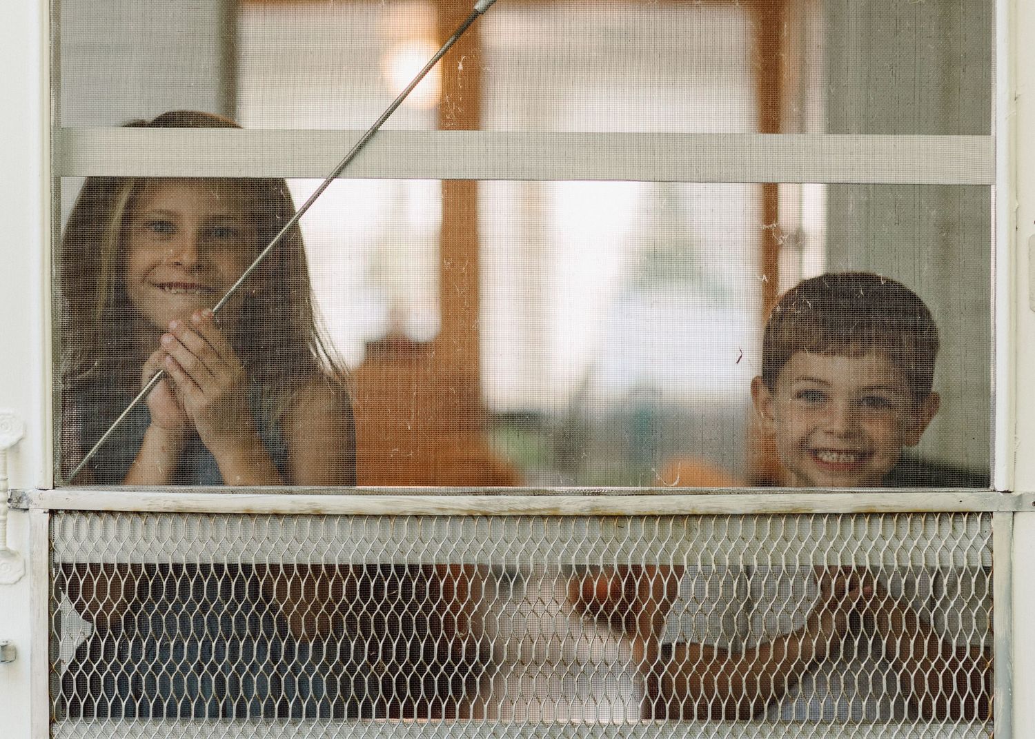 Two children looking through a screened window with happy expressions.