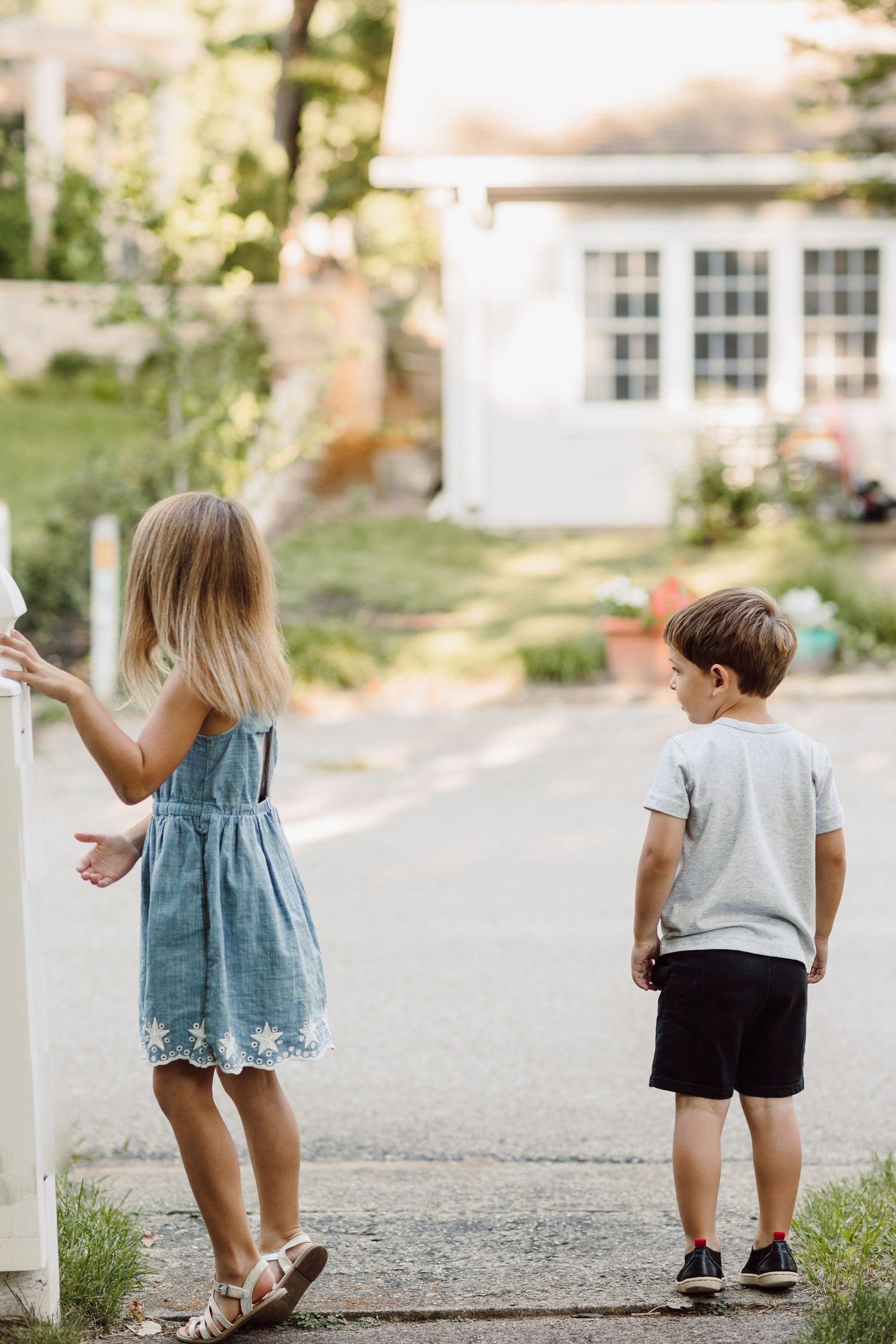 Two young children play together outdoors near a white house on a gravel path.