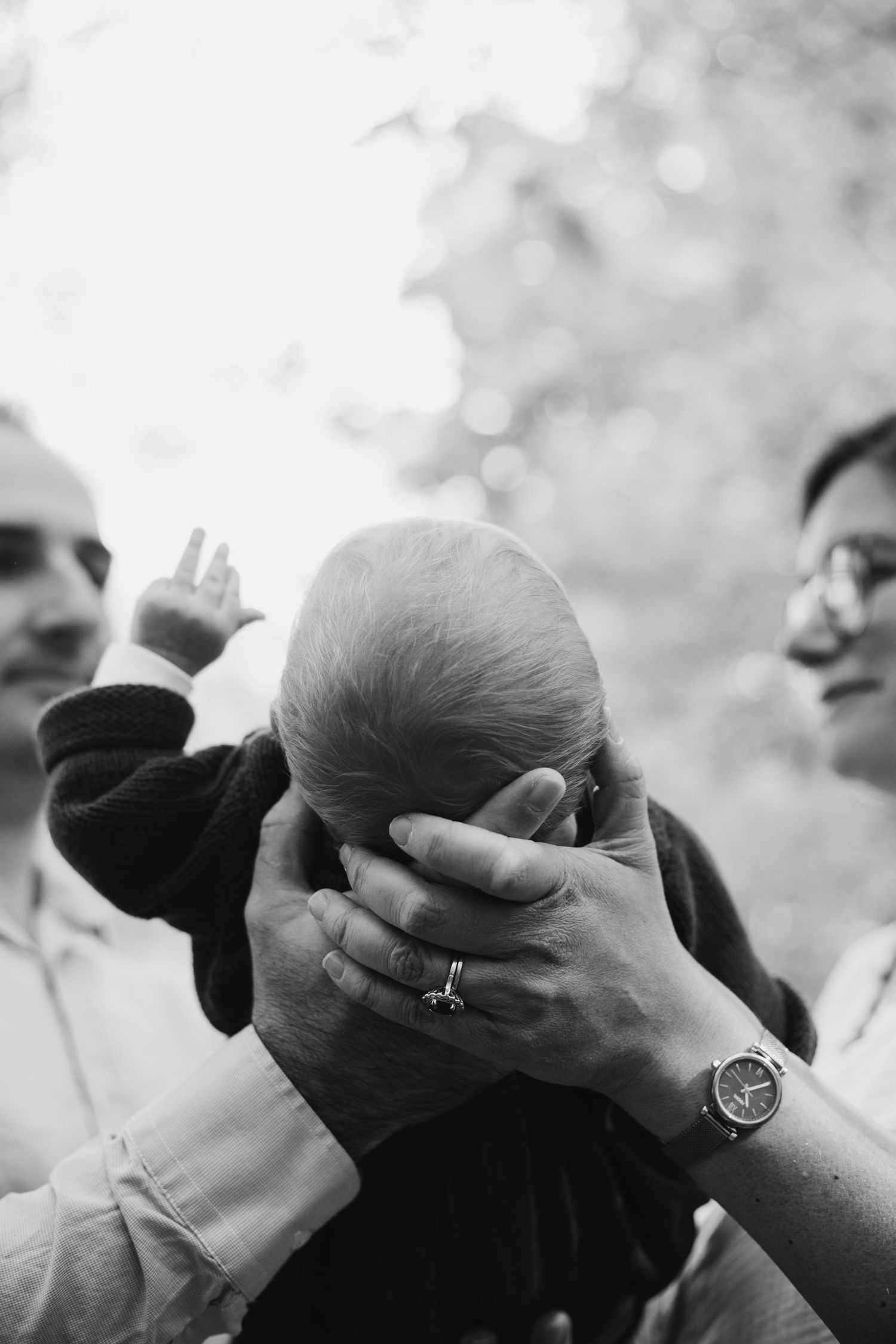 Two adults gently cradling a newborn baby's head outdoors in a black and white photo.