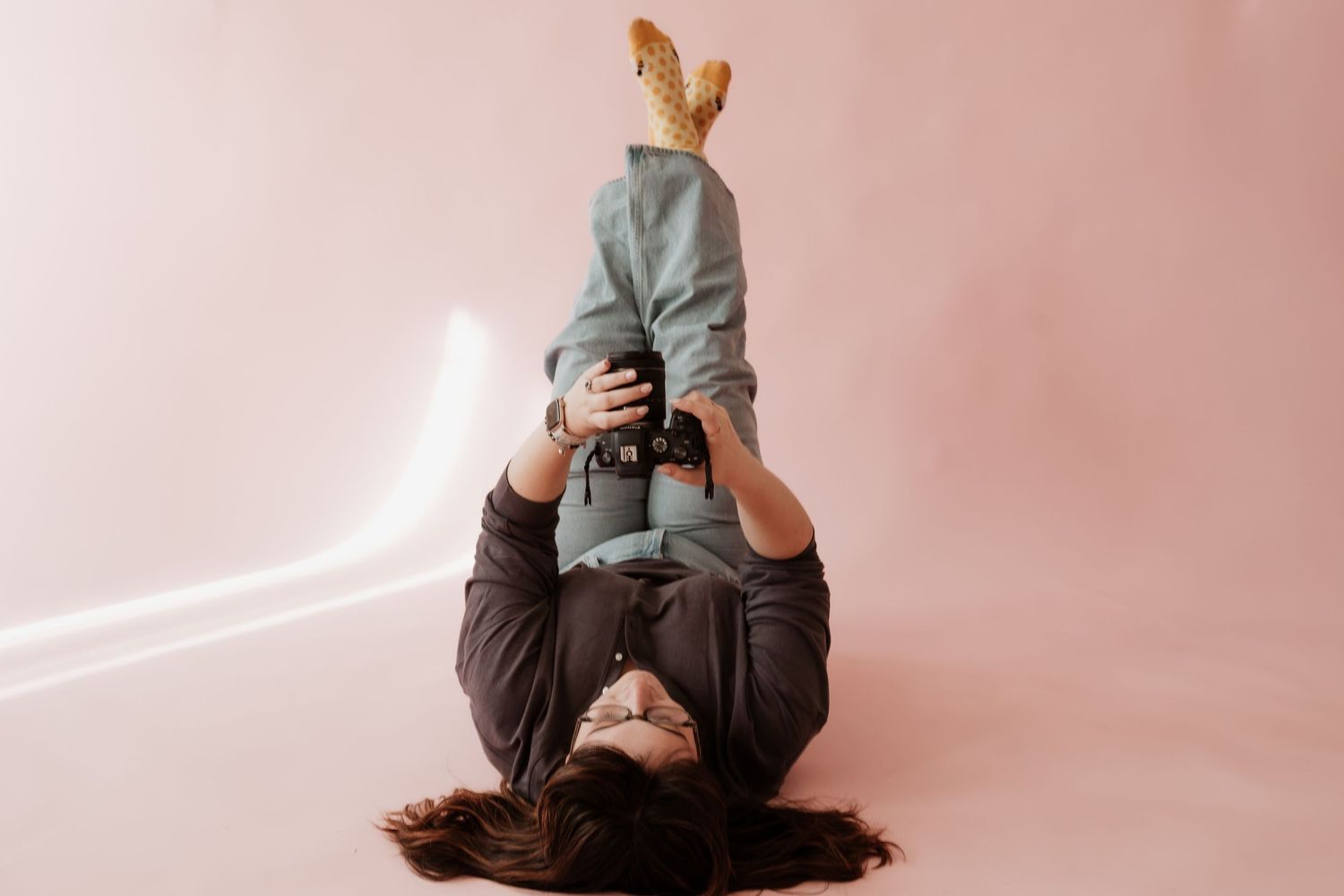 Photographer lying on floor holding camera while taking creative self portrait against pink background.