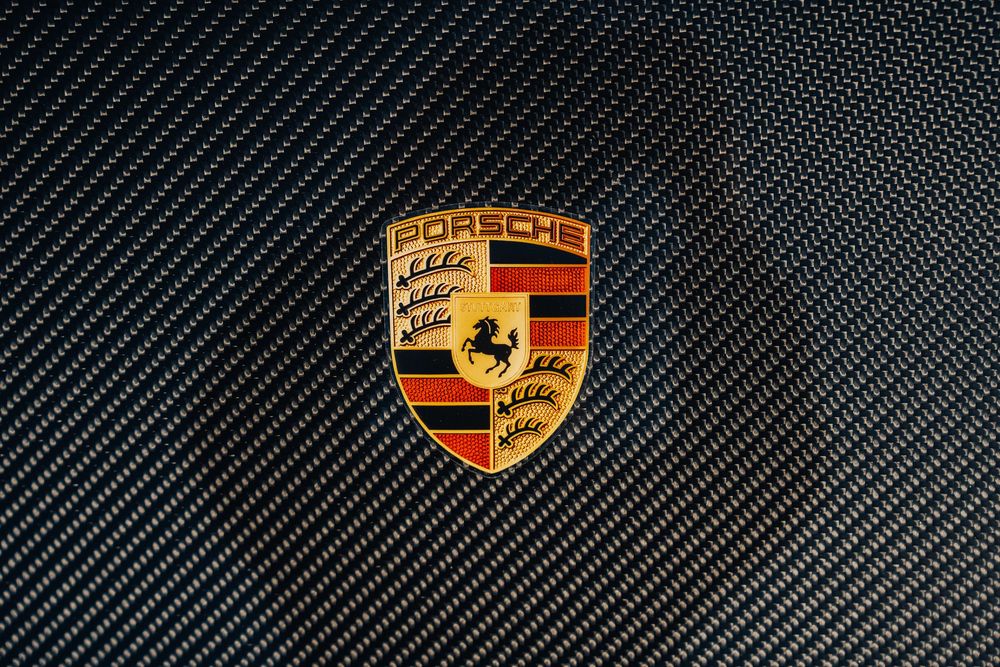 Porsche logo pattern showing seven variations of the iconic crest emblem on a dark carbon fiber background.