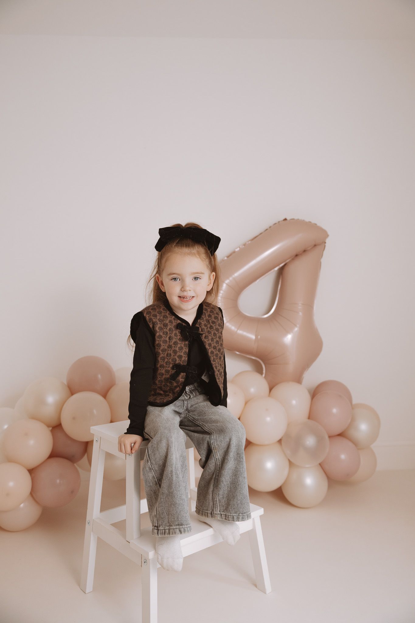 Child in black top and gray pants sits on white chair against pink balloon backdrop in minimalist studio setting.