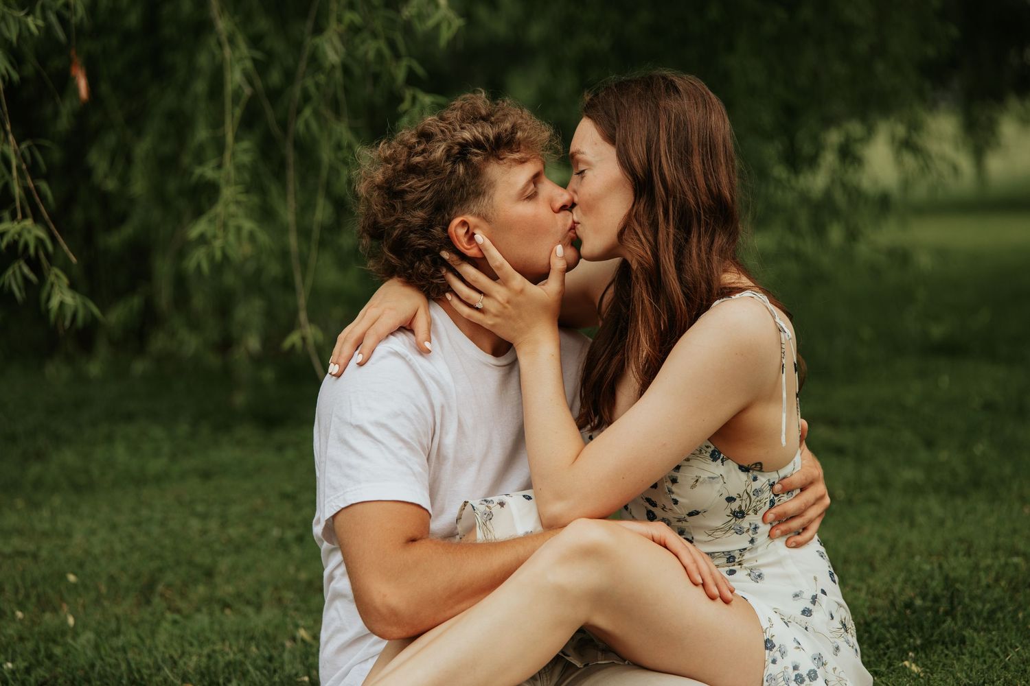 Two people share an intimate kiss in a lush green outdoor garden setting during a romantic photoshoot.
