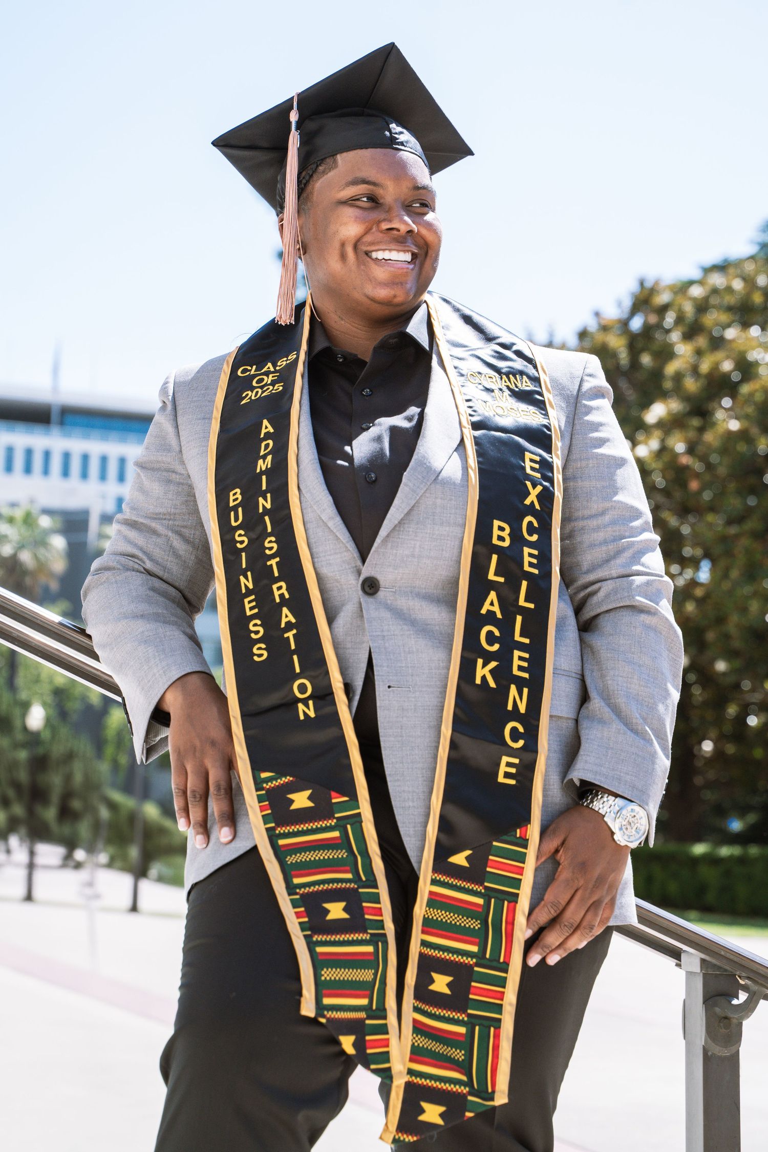 Graduate wearing traditional kente stole and graduation cap poses outdoors in grey suit.