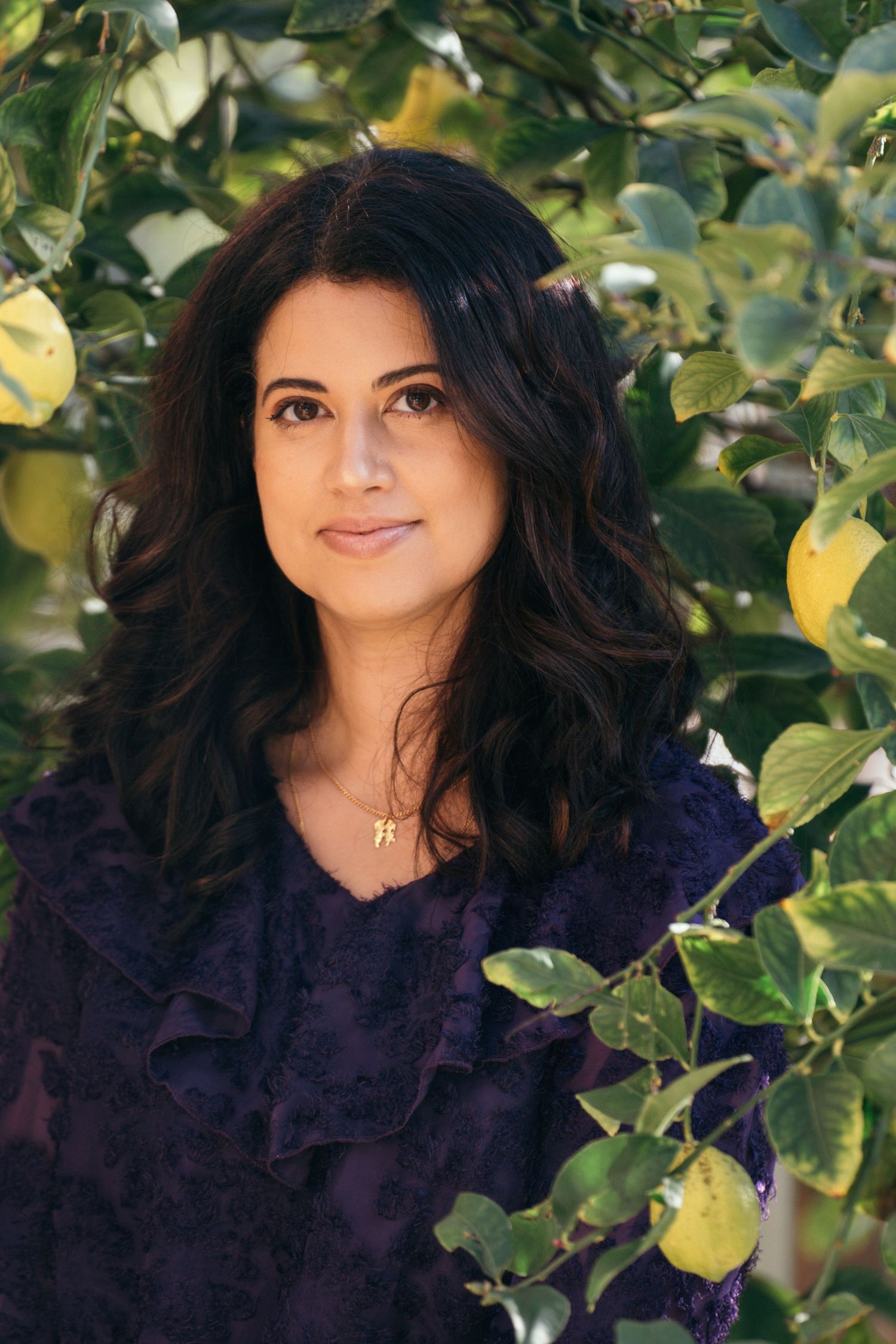 A person with dark hair poses outdoors against green foliage in natural lighting.