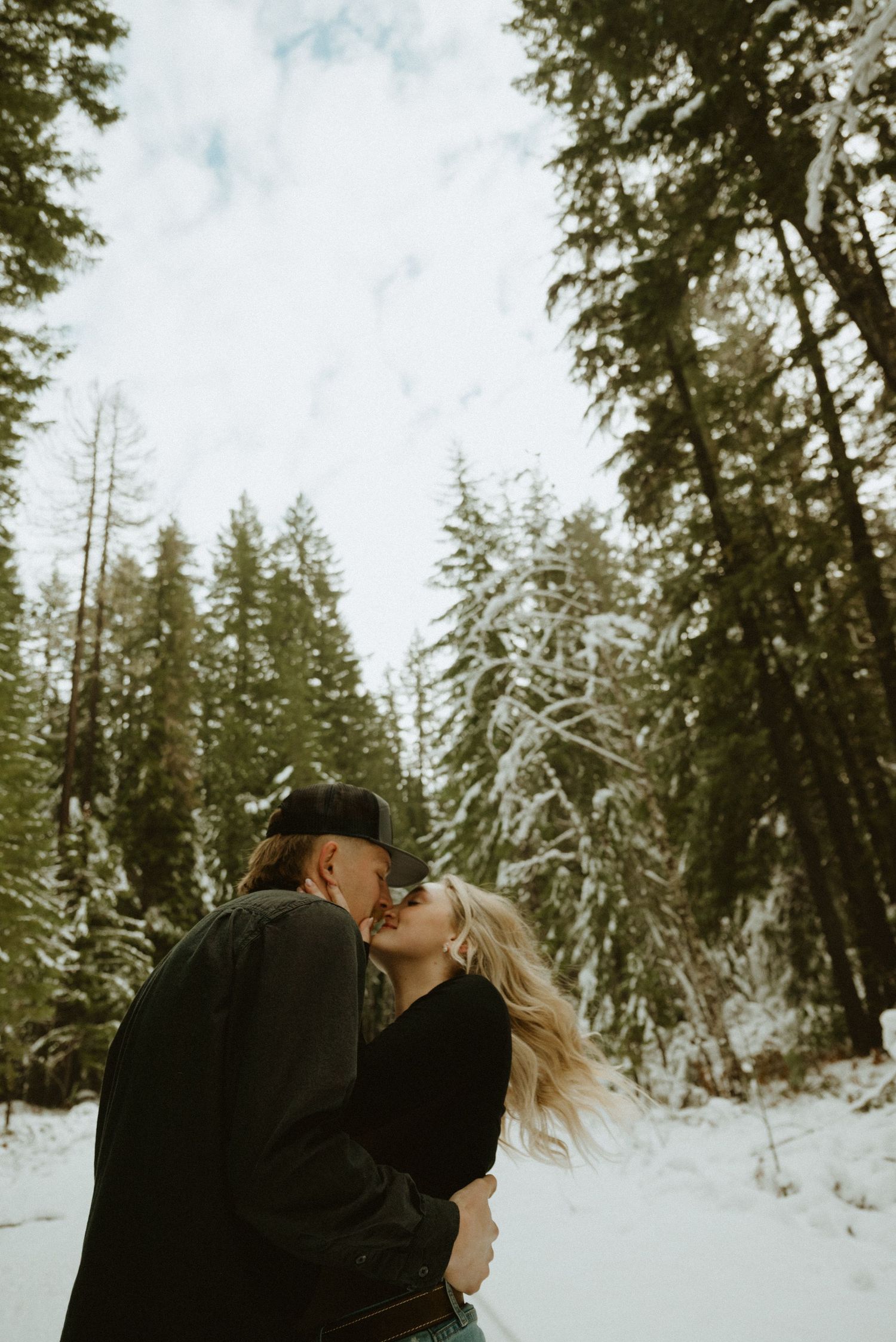 A couple embraces in a snowy winter forest with evergreen trees in the background.