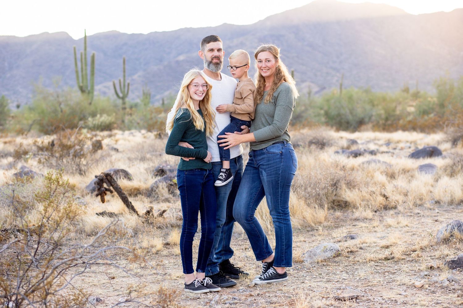 Young family of four smiling together in Arizona desert with saguaro cactus and mountains.