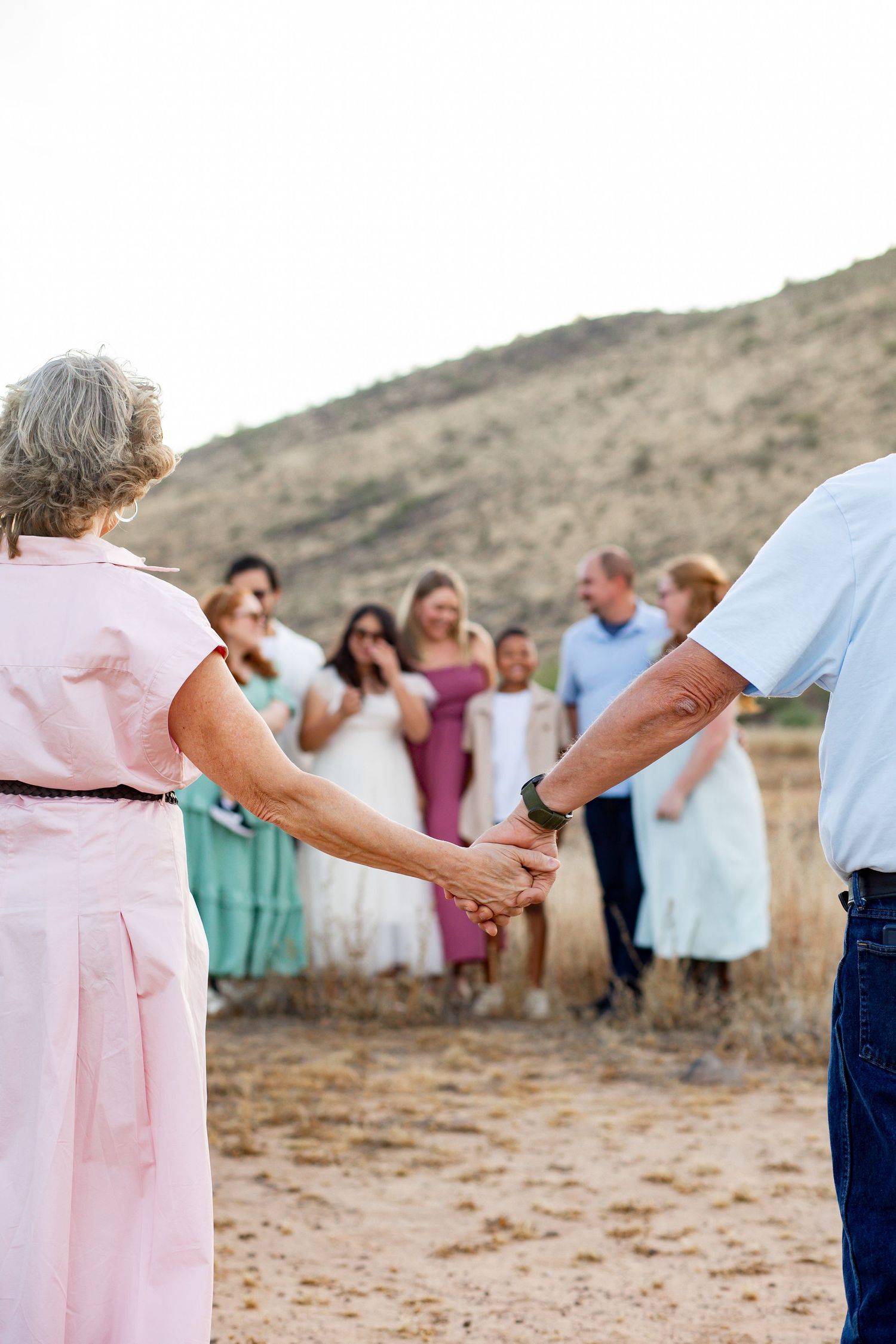 Elderly couple holding hands walking toward a group of family members gathered outdoors in a desert landscape.