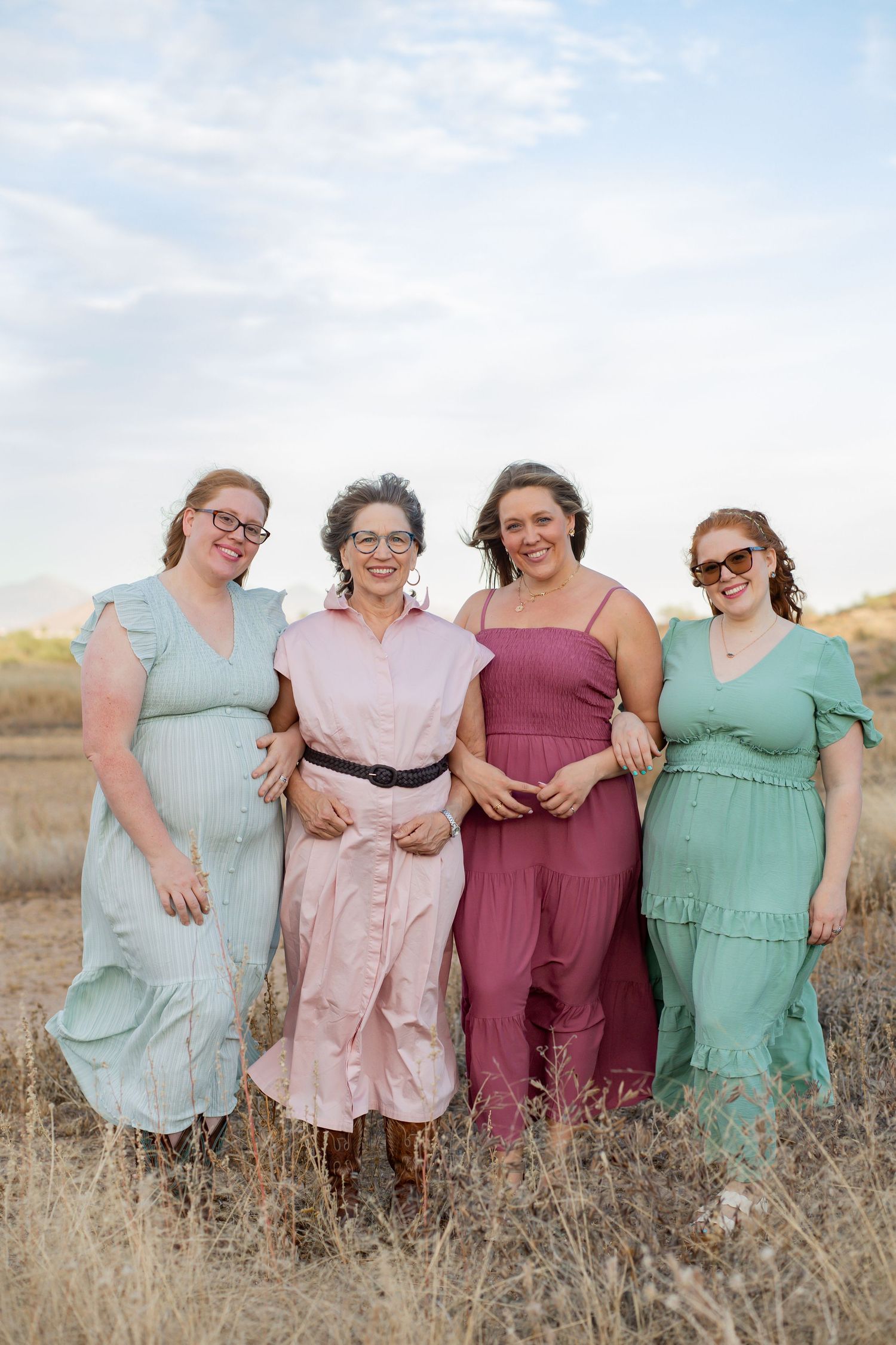 Four women in pastel dresses smile together in a golden field under a cloudy sky.