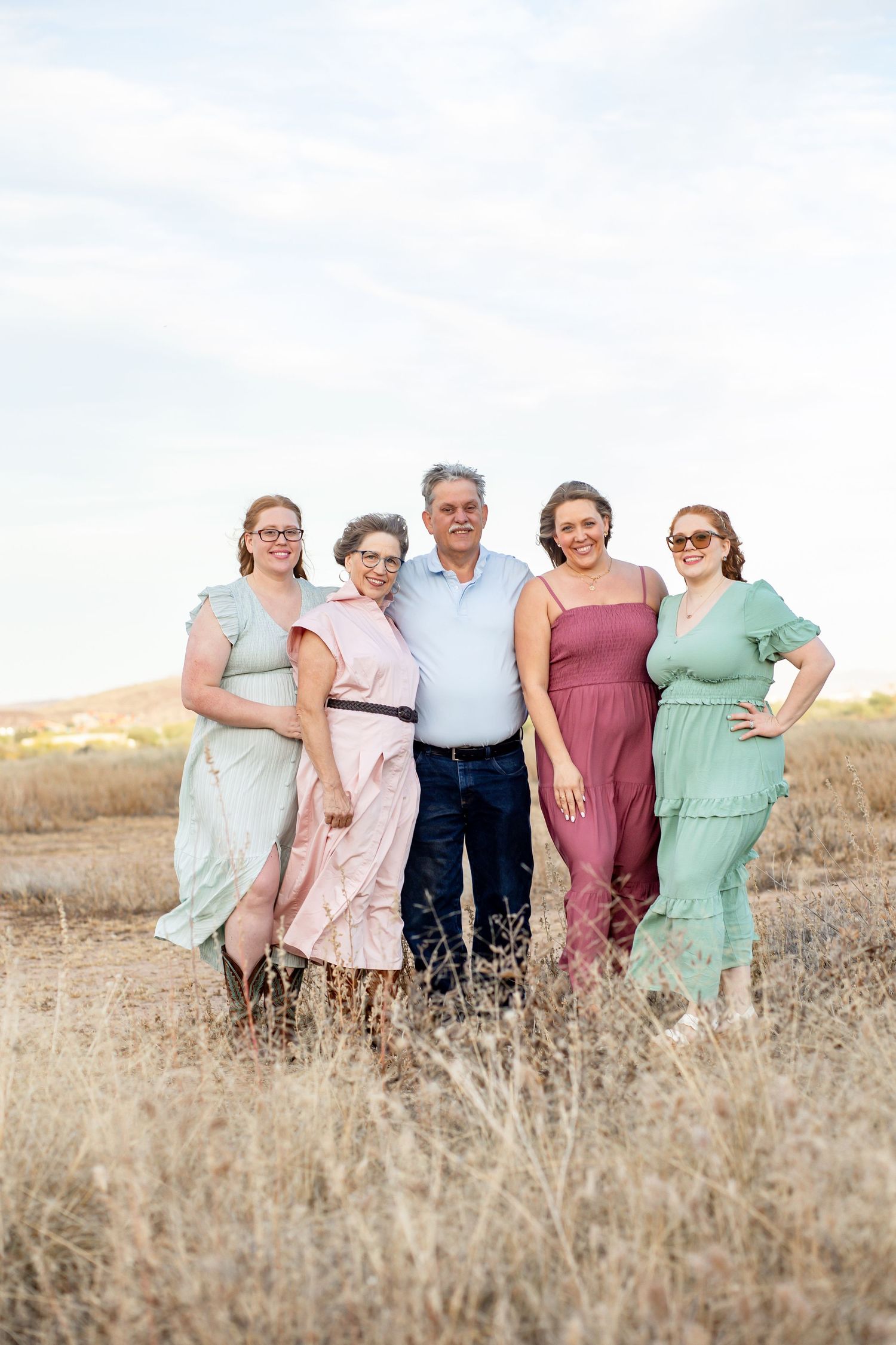 Family of five posing outdoors in a dry field, wearing pastel dresses and casual attire, smiling together under a cloudy sky.