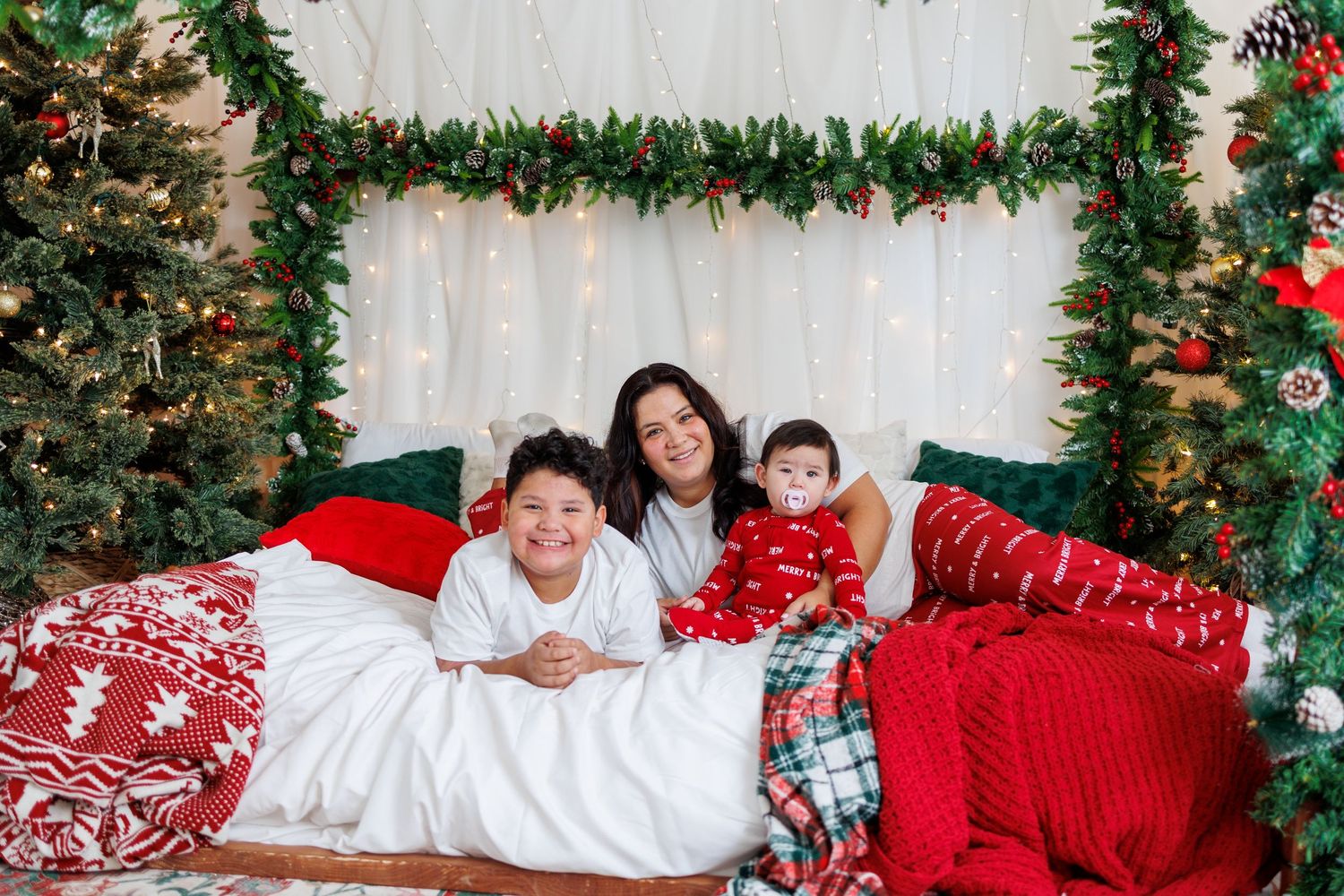 Mother with two children posing on festive Christmas bed with holiday decorations and string lights.