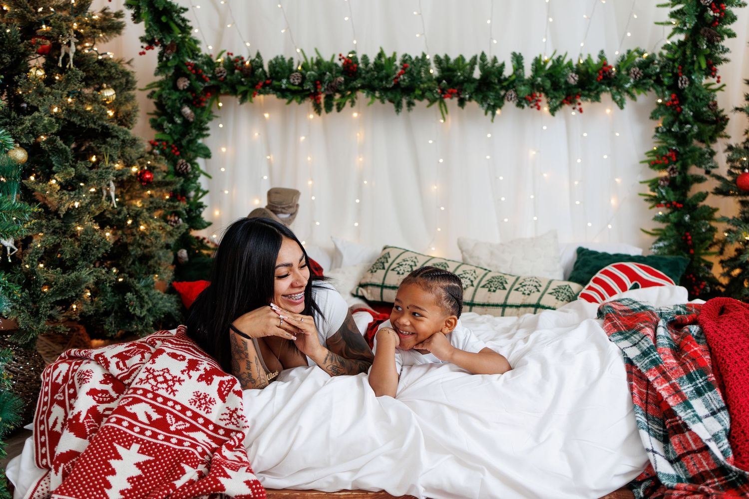 Mother and baby relaxing on festive Christmas bed setup with holiday lights and decorated tree nearby.