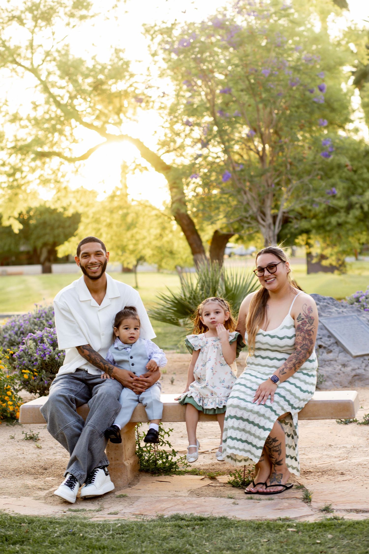 Family of four posing on a stone bench in a sunlit park, parents with two young children, surrounded by lush greenery.