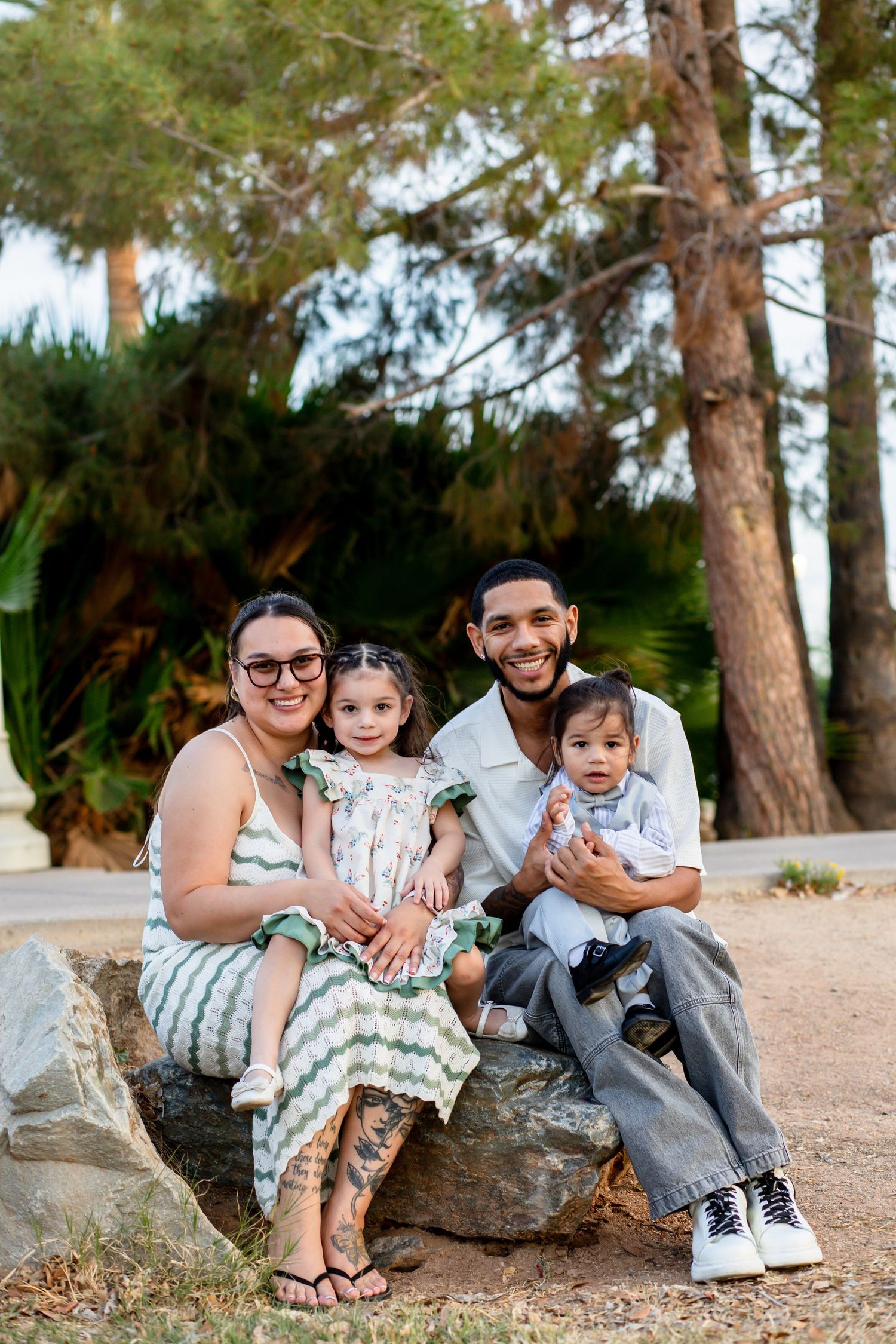 Family of four smiling on a rock outdoors, with two young children, surrounded by lush green trees.